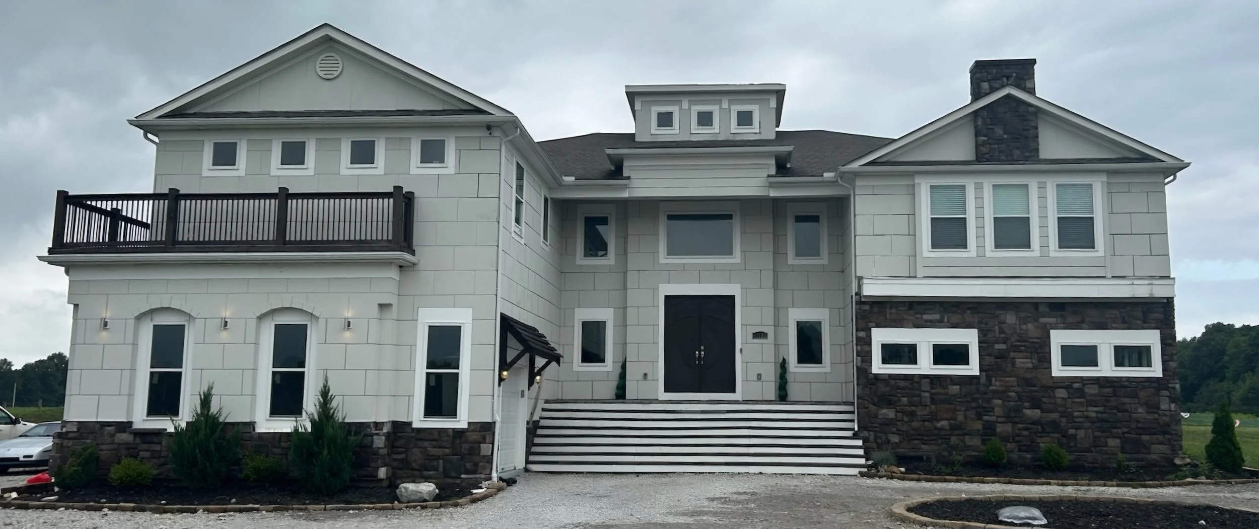 A modern multi-story house with a mix of white and dark stone exterior, multiple windows, and a front staircase leading to a large double door entrance.