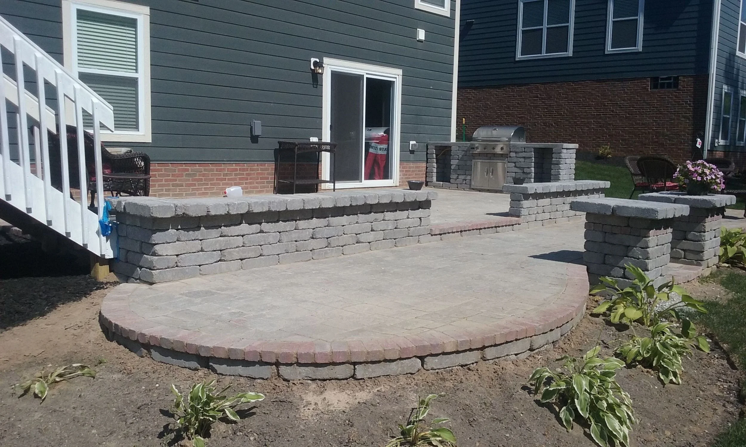 Backyard patio with built-in brick and stone fire pit, outdoor seating, and a built-in stainless steel grill, with a house in the background.
