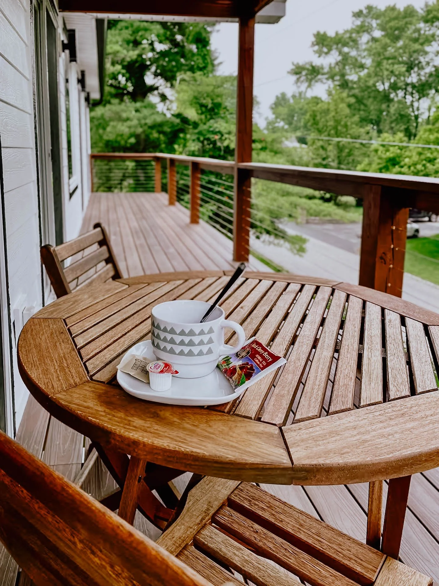 A wooden patio table with a cup, small container, and packet of strawberry jelly, set on a balcony with a view of trees and a street.
