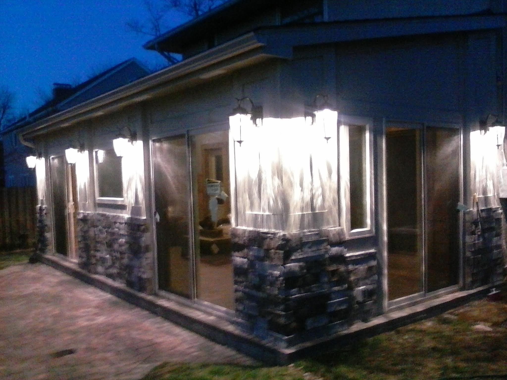 A house under construction or renovation, covered with plastic sheeting and surrounded by rocks, with a stone foundation and large windows. The photo is taken during dusk or early evening.
