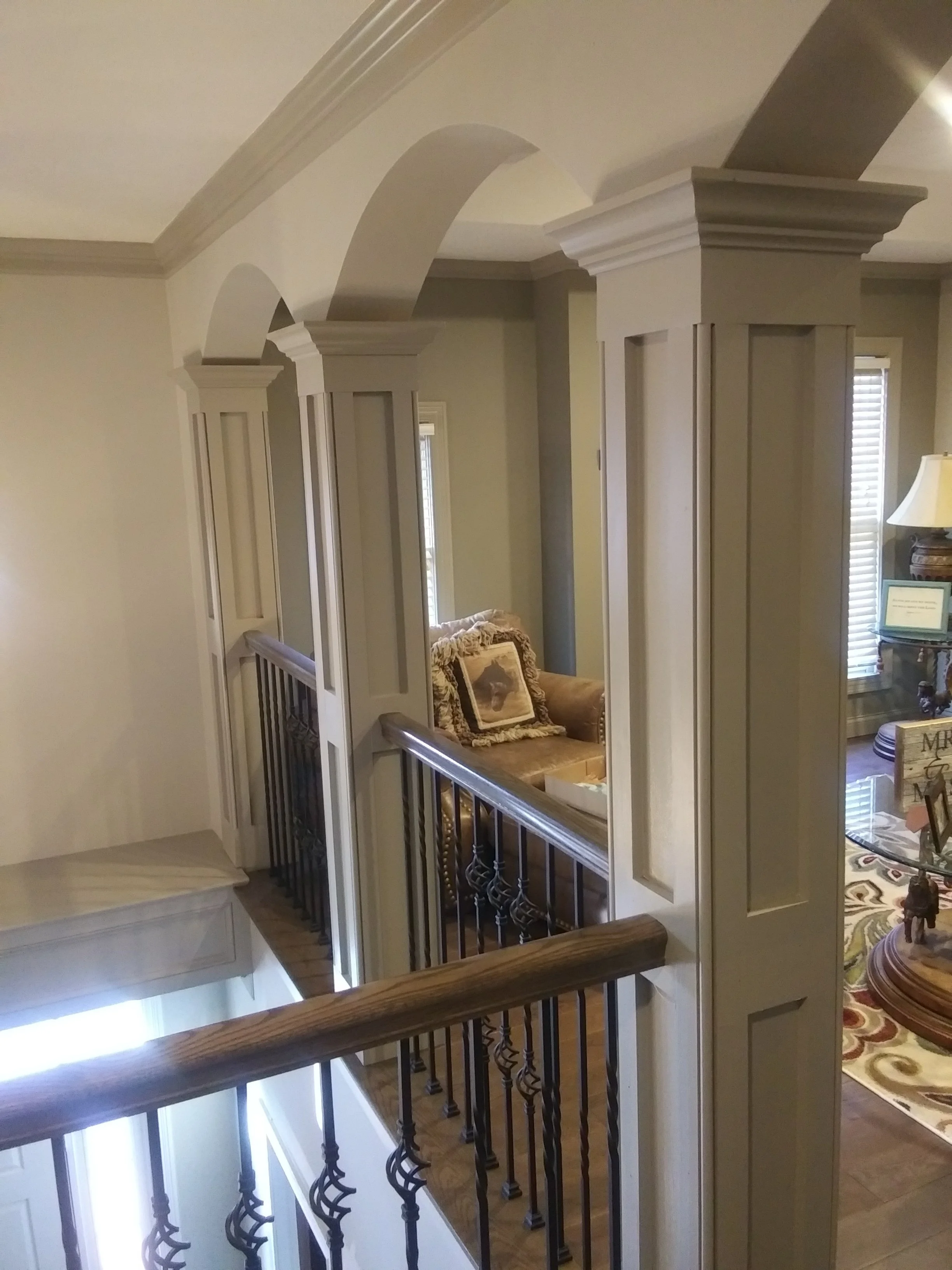 Interior view of a house showing a staircase with a wooden handrail and decorative iron balusters, overlooking a living area with furniture and windows with blinds.