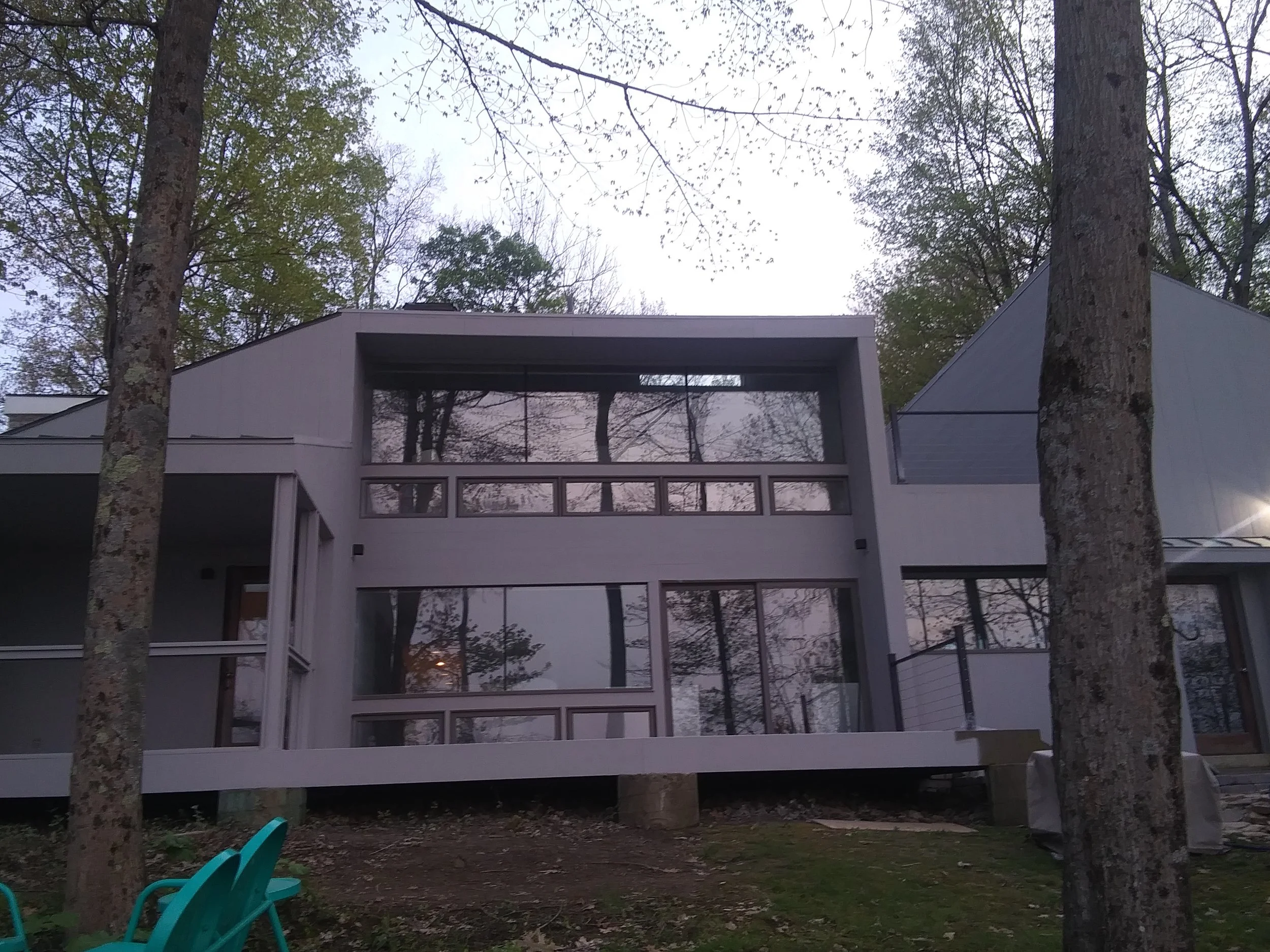 View of a modern house with large windows, surrounded by tall trees with green and bare branches, showing a partly cloudy sky at dusk.