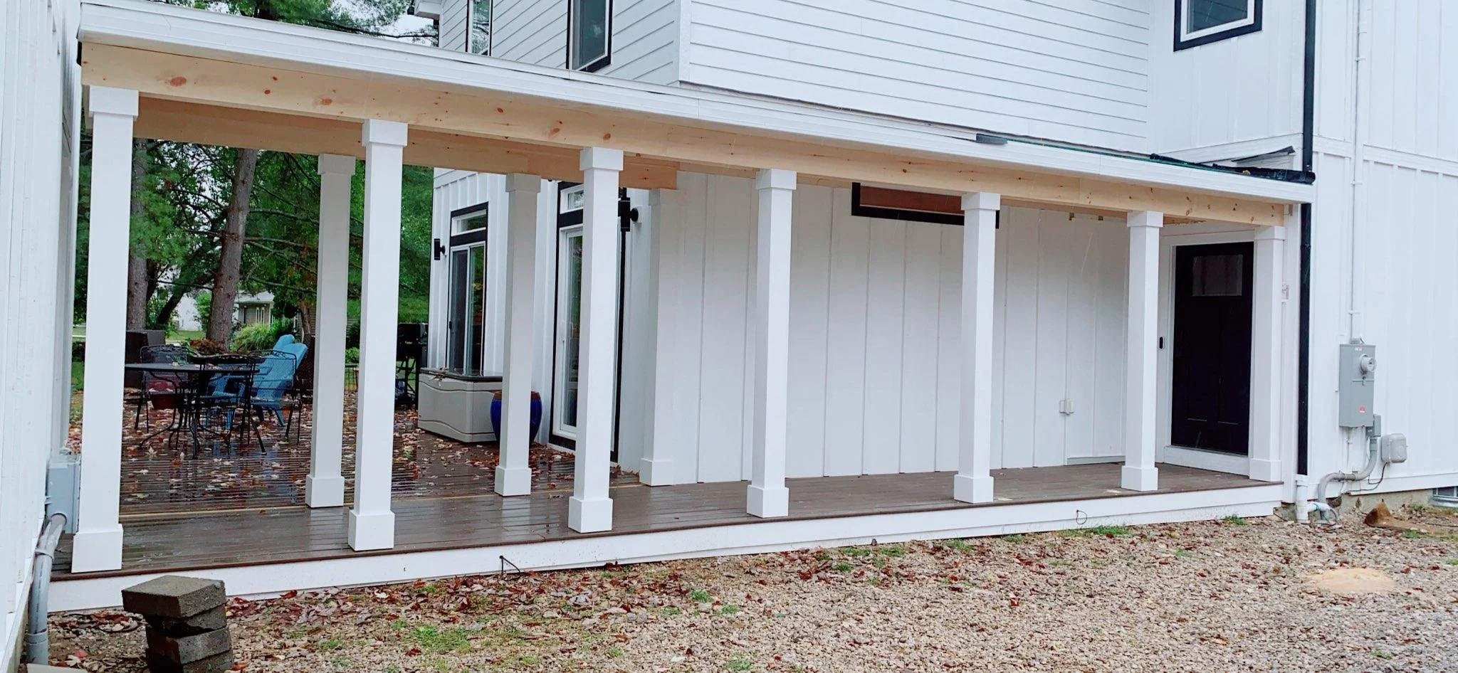 Back porch of a white house with a new wooden deck and white support columns, outdoor furniture, and trees in the background.