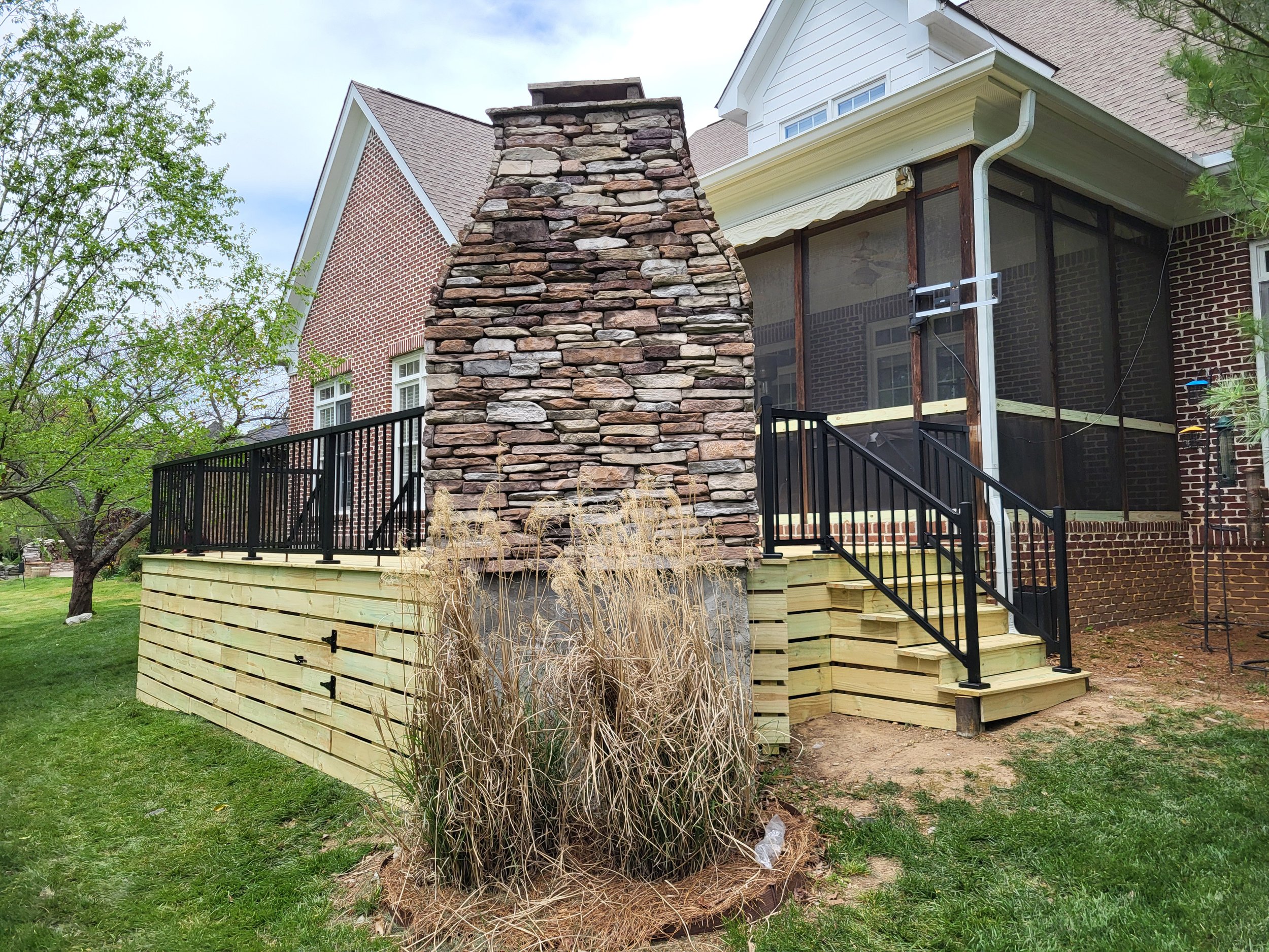 A house with brick exterior, white siding, and a porch under construction. There is a stone chimney and a black metal railing around the porch.
