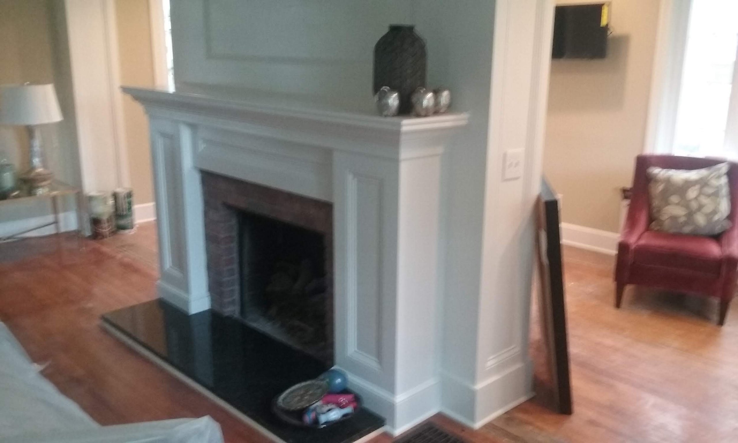 Photo of a living room interior featuring a white fireplace with a black interior, decorated with a black vase and silver decorative items on top. To the left, there is a side table with a white lamp and candles. To the right, a red armchair with a patterned cushion is visible, with a window and part of another room in the background.