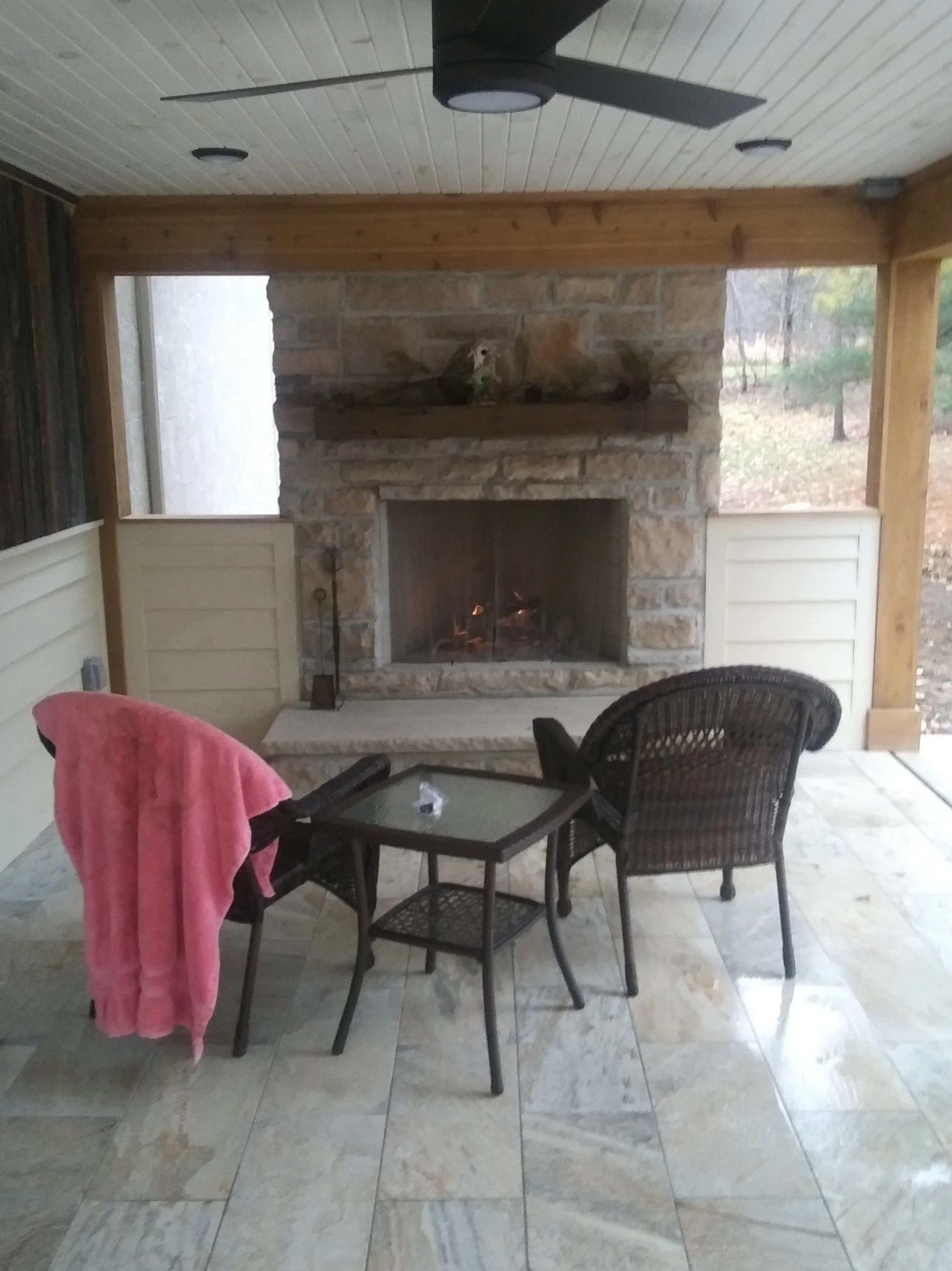 Outdoor patio area with a fireplace, wicker chairs, and a glass-top table. A pink towel is draped over one chair. The patio has a tiled floor and a screened enclosure with a view of trees outside.