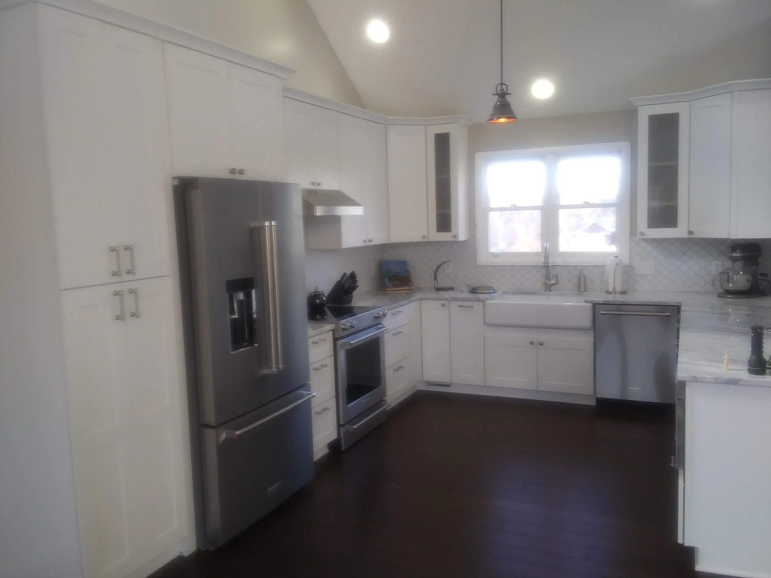 A kitchen with white cabinets, a stainless steel refrigerator, a stove, a sink under a window, and pendant lighting, with dark hardwood floors.