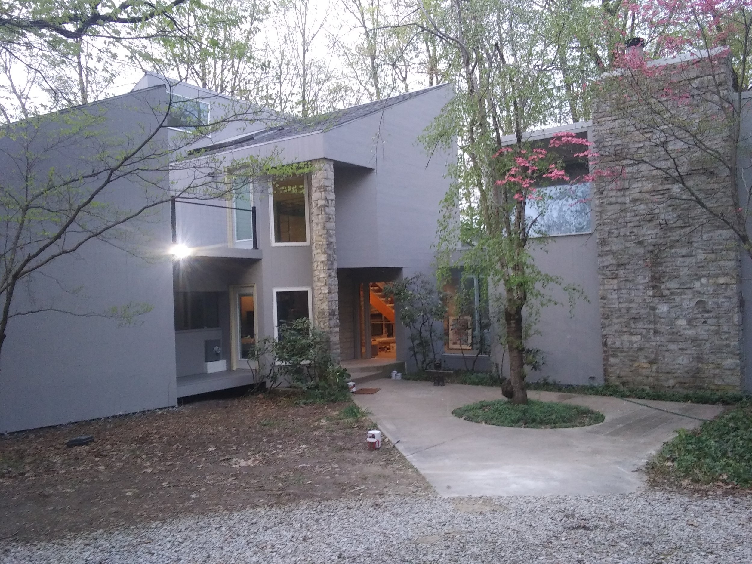 Photo of a modern multi-story house with gray walls, large windows, and a stone accent wall. There are trees with budding leaves and pink flowers surrounding the house, and a concrete walkway leads to the entrance. The scene appears to be taken at dusk or early evening.