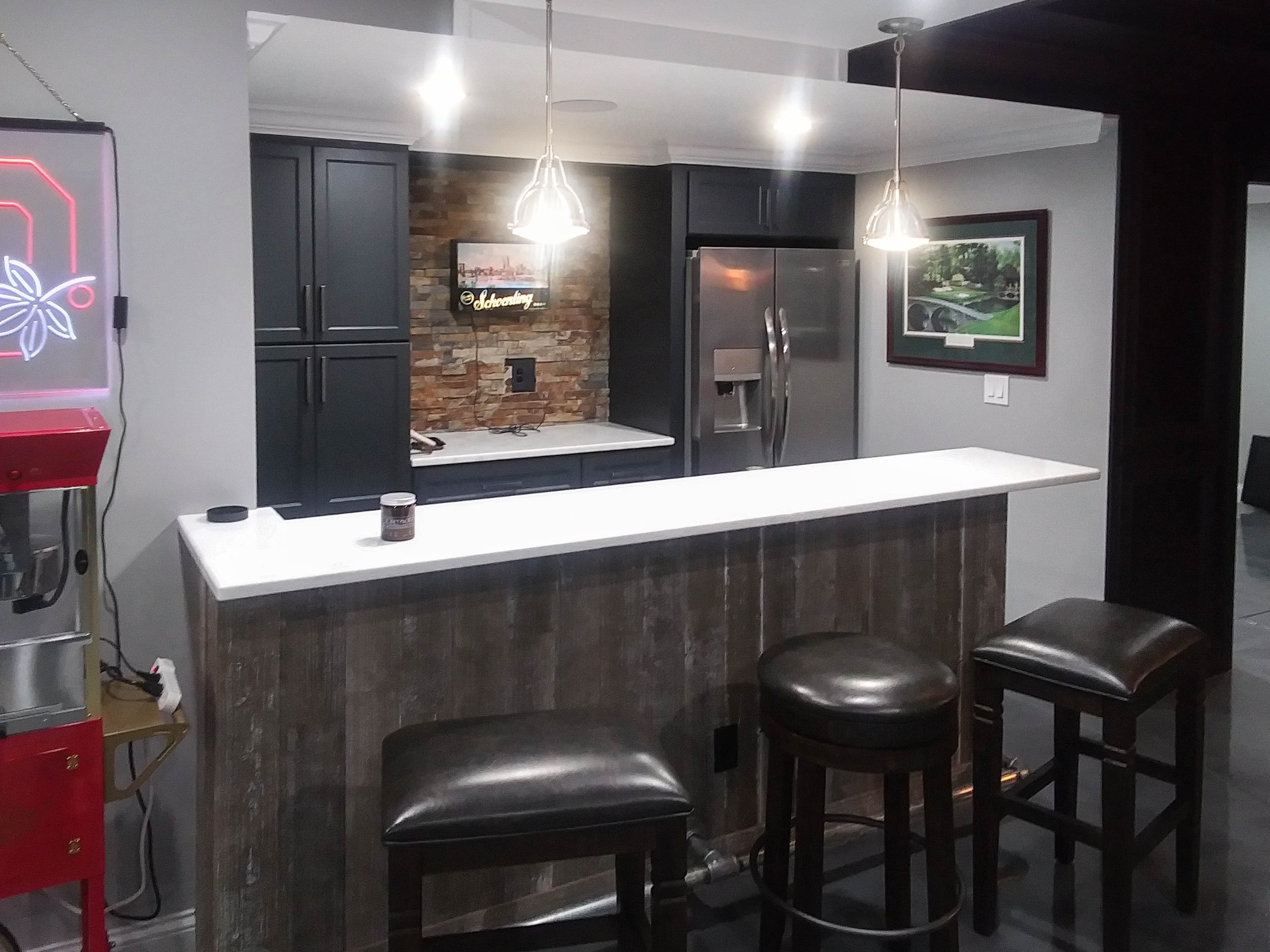 Interior view of a modern kitchen with dark cabinets, a stone accent wall, stainless steel refrigerator, pendant lights, and a white countertop with stools.