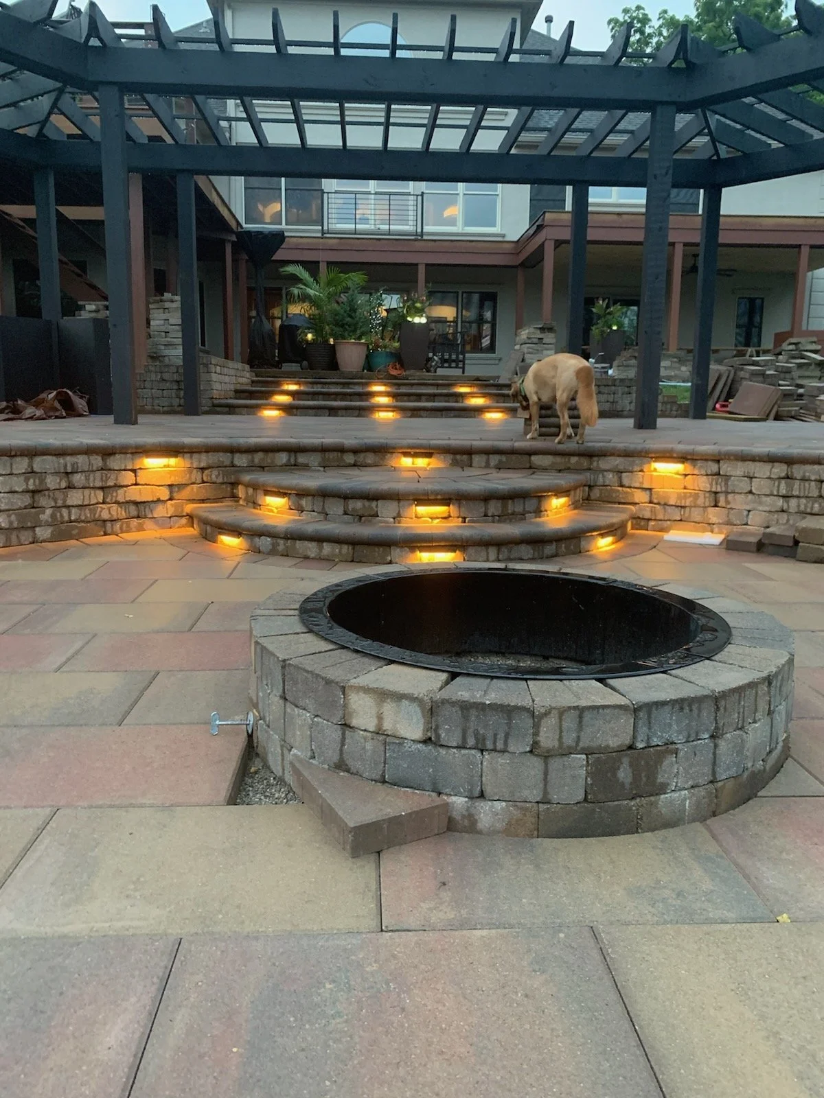 Backyard with brick steps illuminated by embedded lights, a fire pit in the foreground, and a dog sniffing near the steps. There is a covered patio area with potted plants and a modern house in the background.