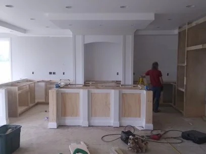 Kitchen under construction with light wood cabinets and a central island, while a worker in a red shirt is working in the background.