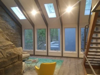 Interior of a living room with large windows and skylights, a yellow chair, a blue rug, and a stone fireplace.