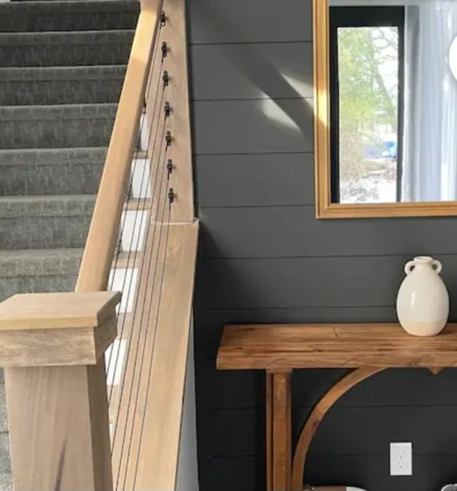 Part of a staircase with gray carpet, adjacent to a gray paneled wall with a window, a wooden table, and a white ceramic vase.