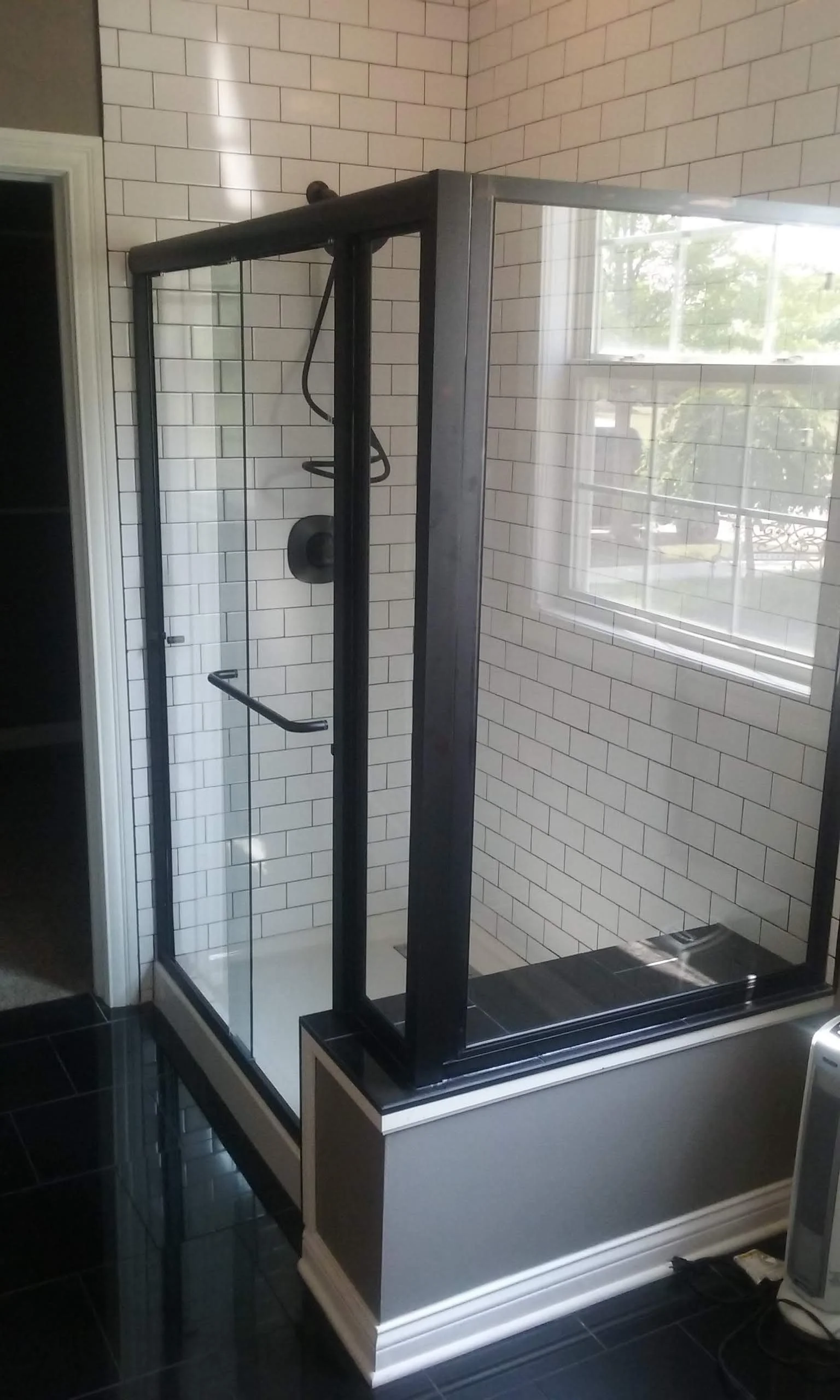 A modern glass-enclosed shower with black framing, white subway tile walls, and a window outside the shower letting in natural light.