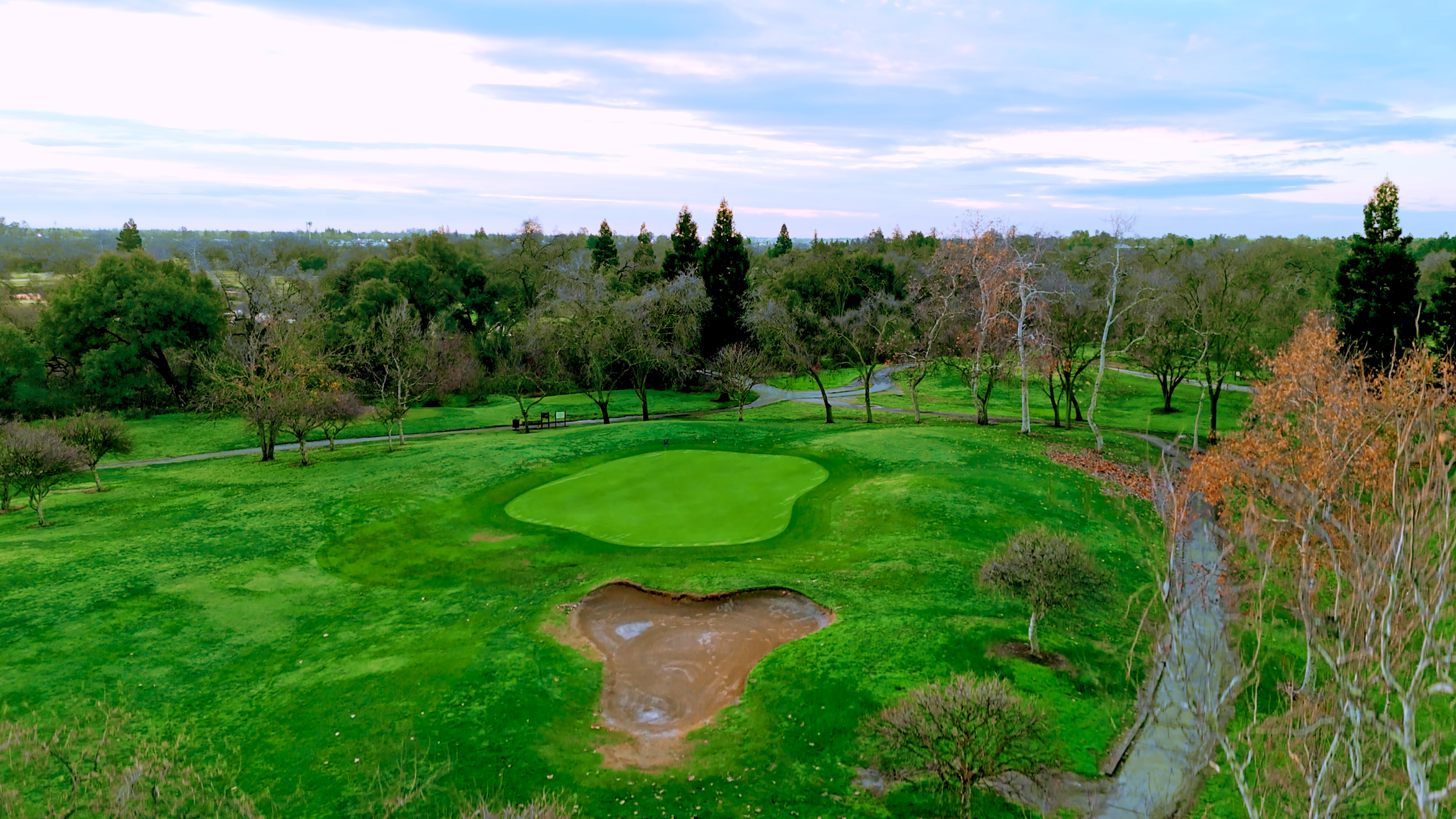 A landscape of a golf course with a green, sand trap, trees, and a creek under a partly cloudy sky.