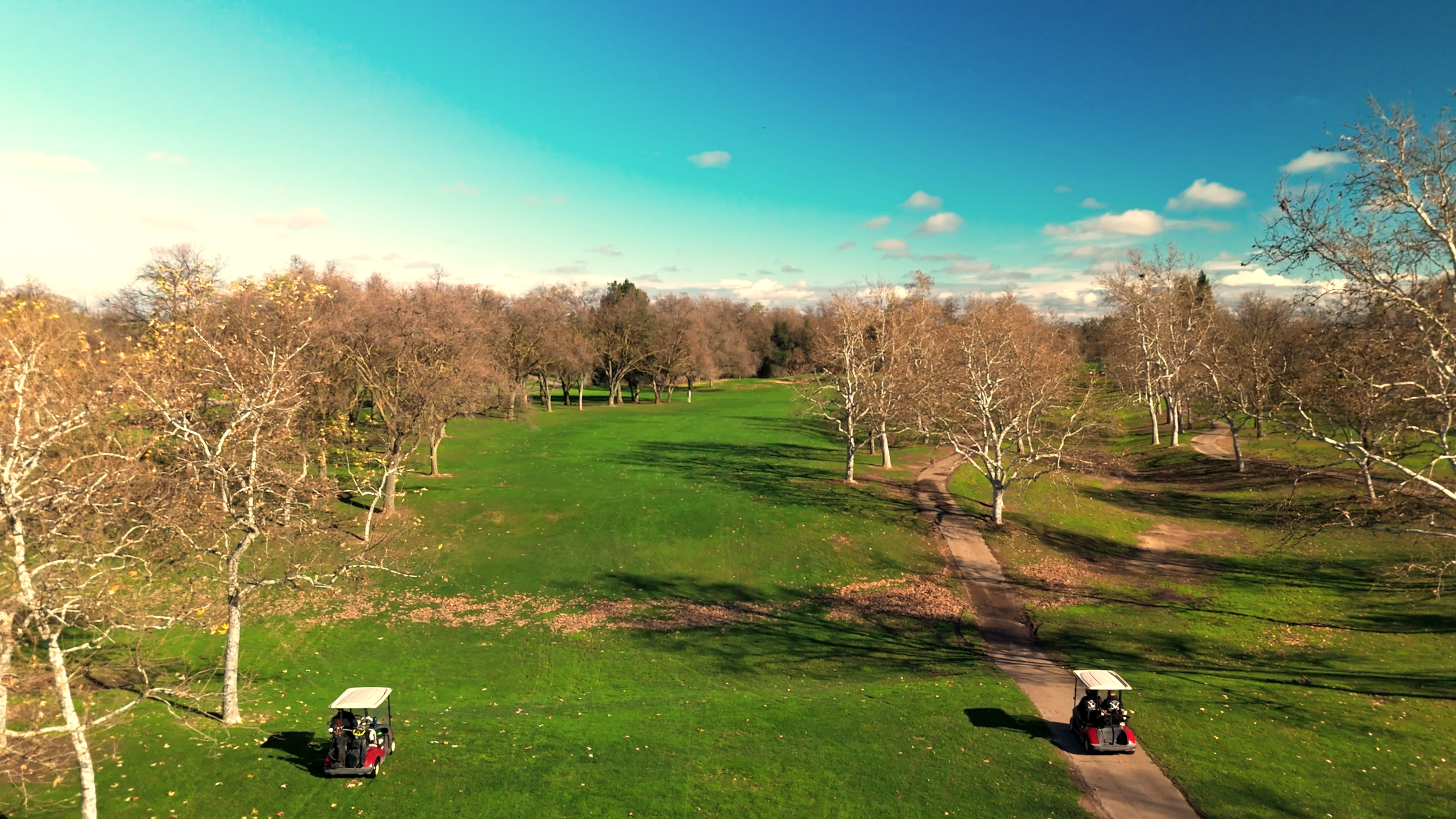 Two golf carts driving on a winding path through a golf course surrounded by leafless trees and a partly cloudy sky.