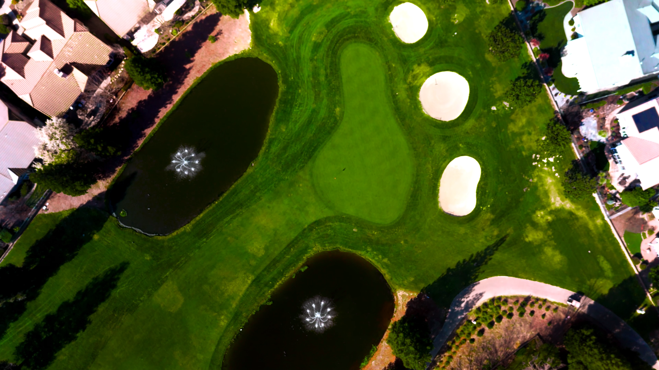 An aerial view of a golf course with green fairways, sand bunkers, two small ponds with fountains, and surrounding residential houses and trees.