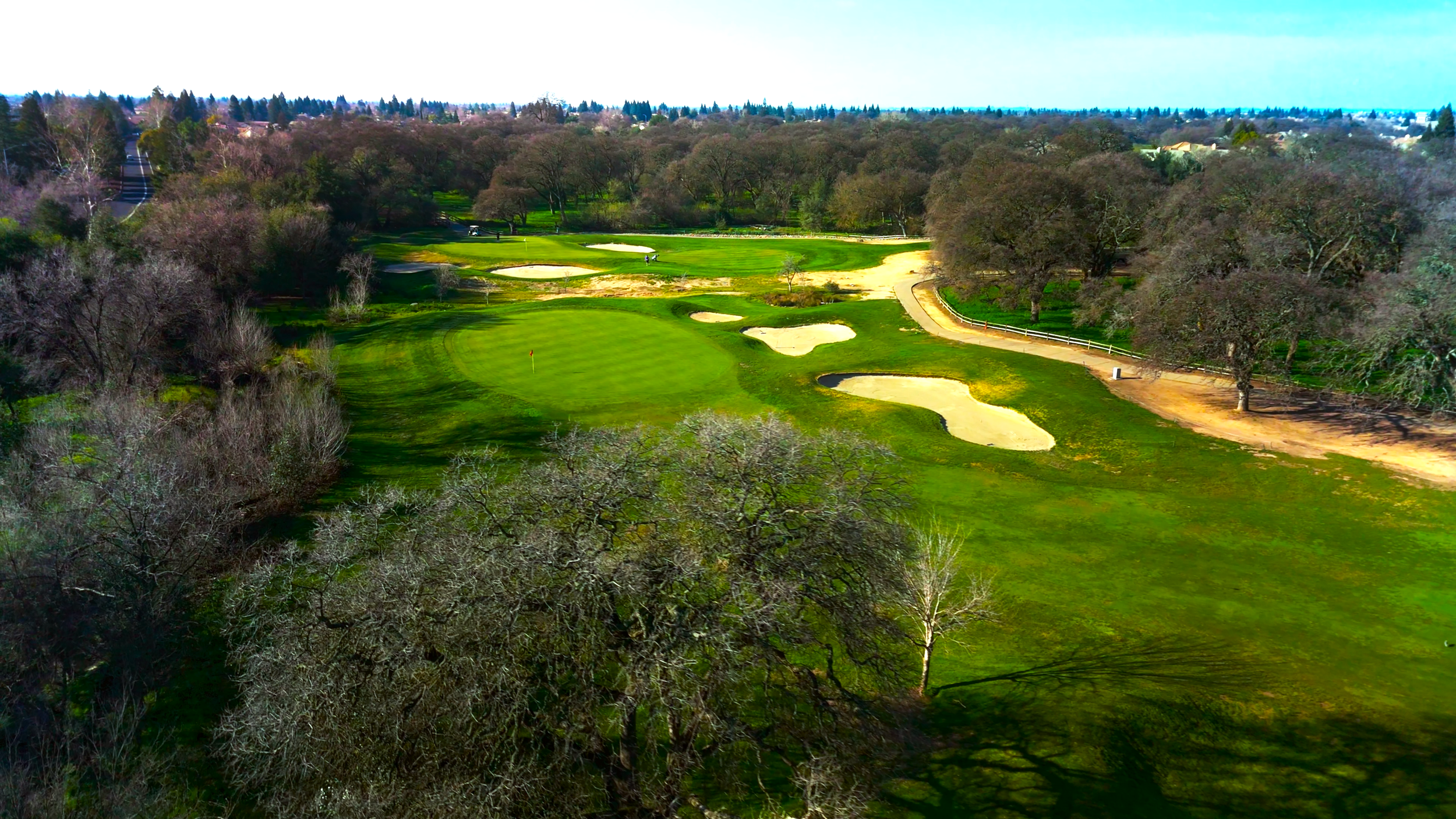 A golf course with green fairways, sand bunkers, and trees under a partly cloudy sky.