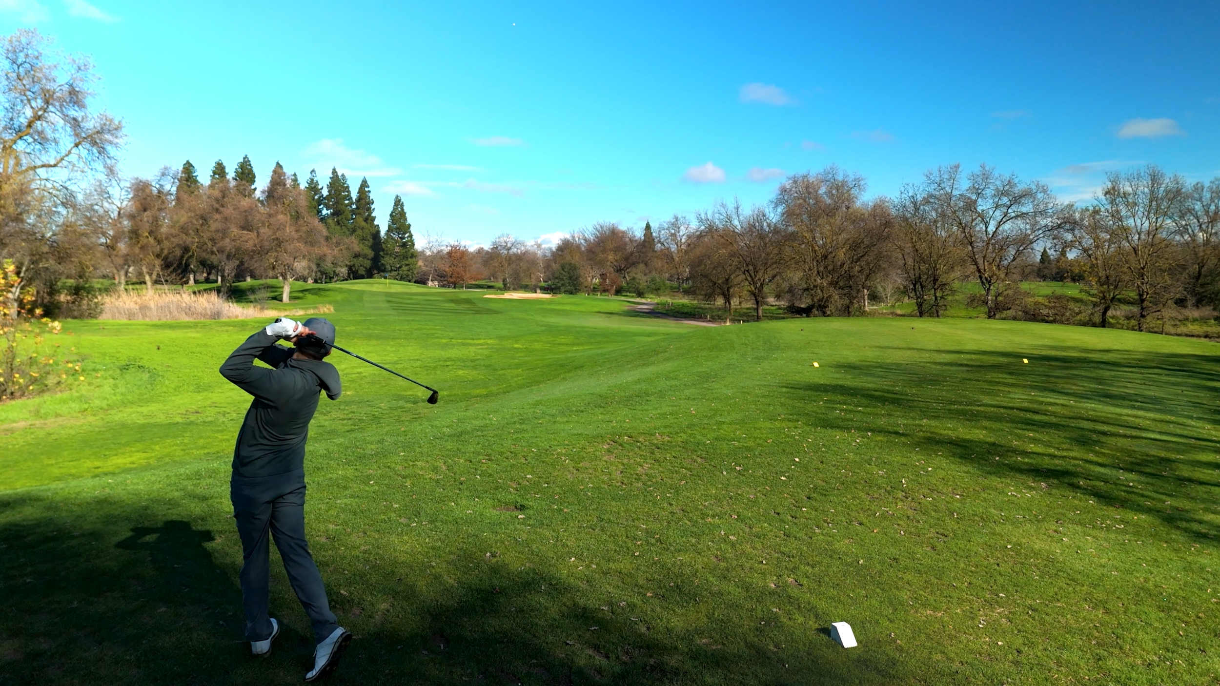 A person in black clothing and a black hat swinging a golf club on a green golf course with trees and a blue sky in the background.