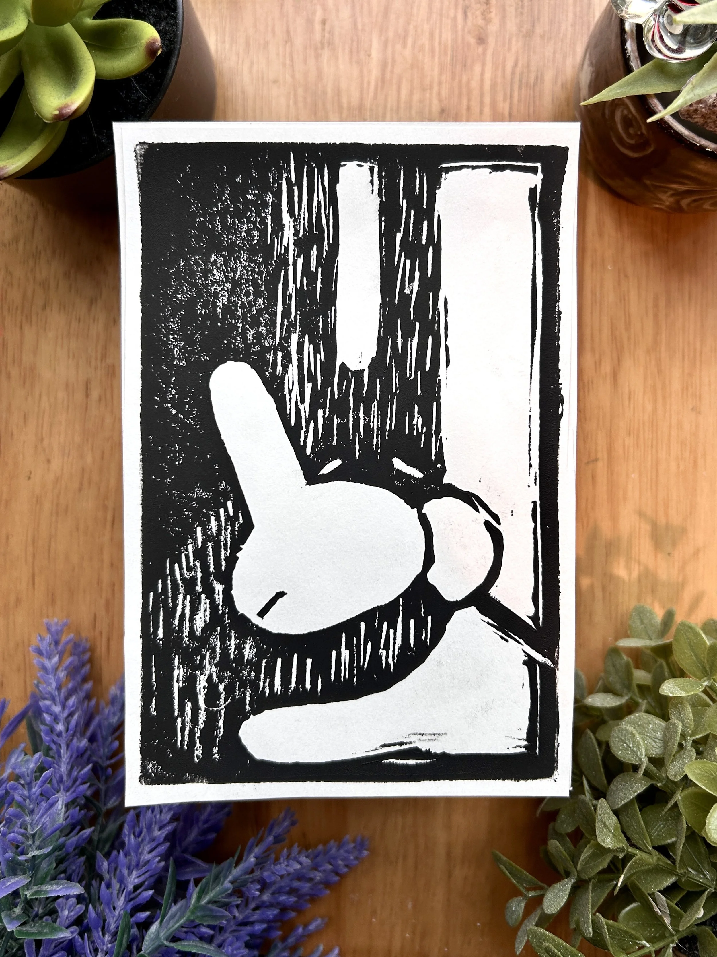 Black and white illustration of a hand making a peace sign with the index and middle fingers raised and the thumb, ring, and little fingers folded down, set against a wood background with plants around.