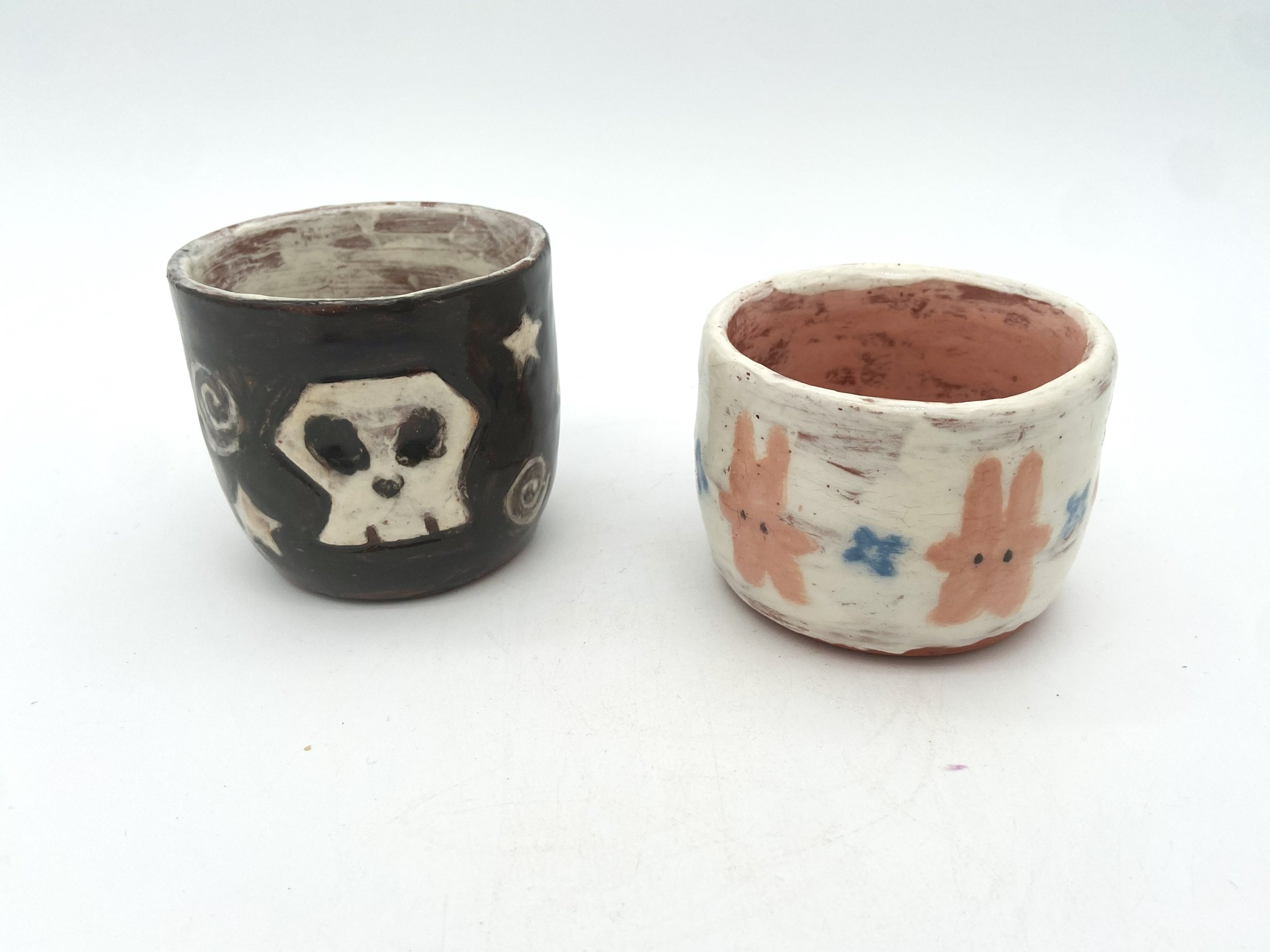 Two small, handmade ceramic cups with painted designs, one featuring a skull with a black background and the other decorated with pink rabbits and blue flowers on a white background.