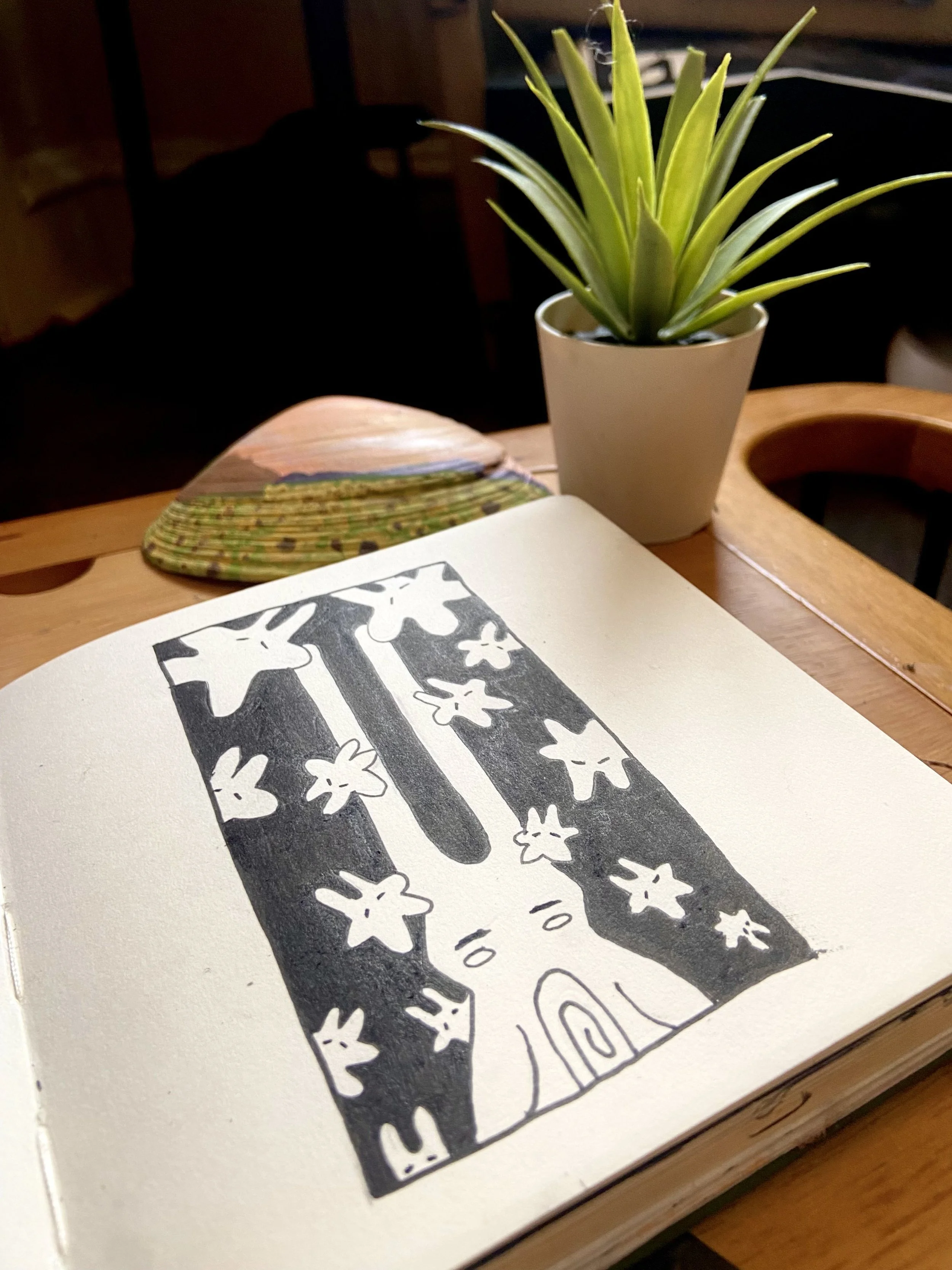 A sketchbook open on a wooden table, featuring a black-and-white drawing of a tree with smiling stars and a rabbit at the base. A potted succulent plant and a colorful, patterned item are also on the table.