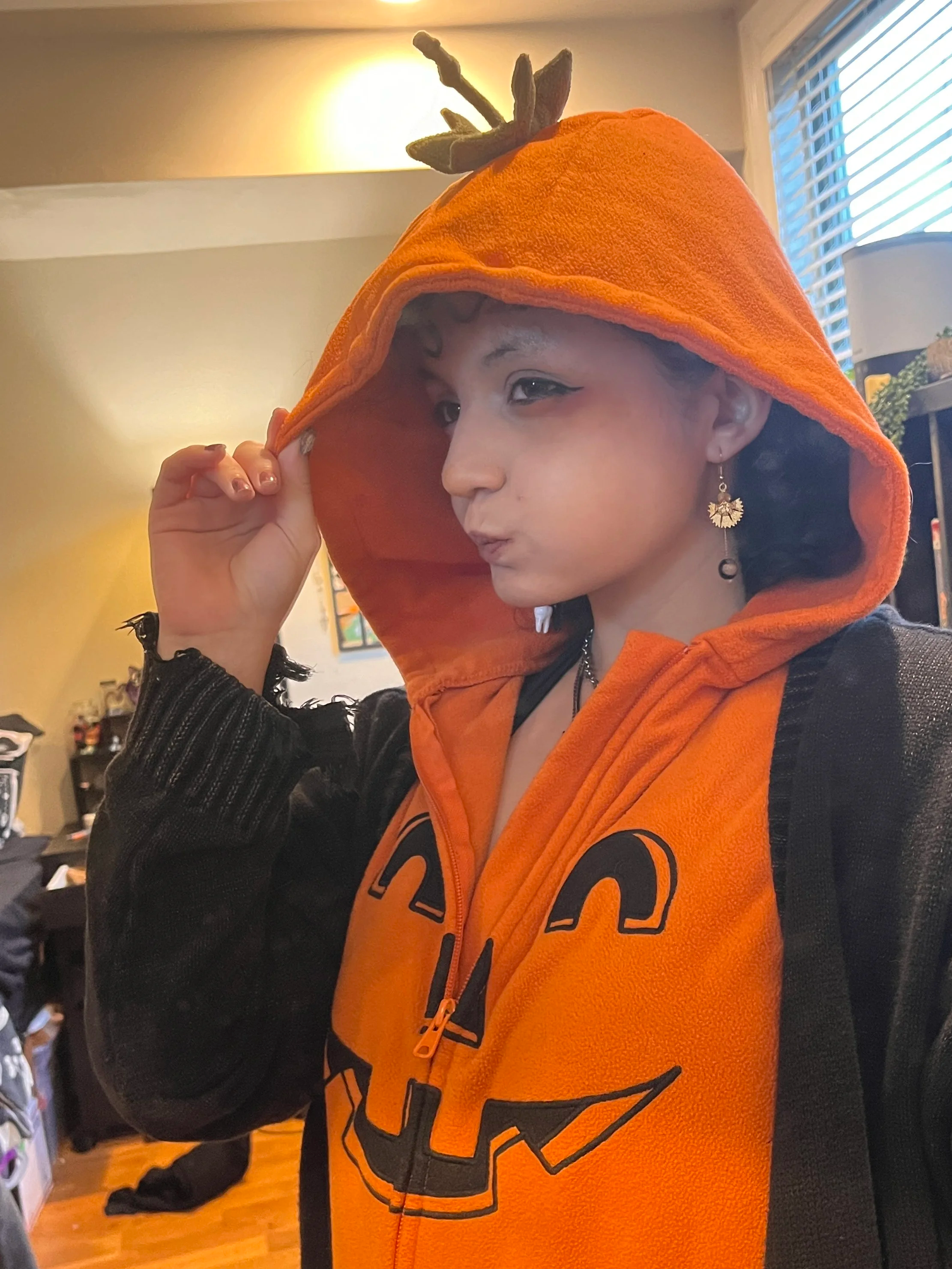 Person wearing an orange pumpkin hoodie with a pumpkin face design, holding the hood with one hand, in a room with a window and a lamp.