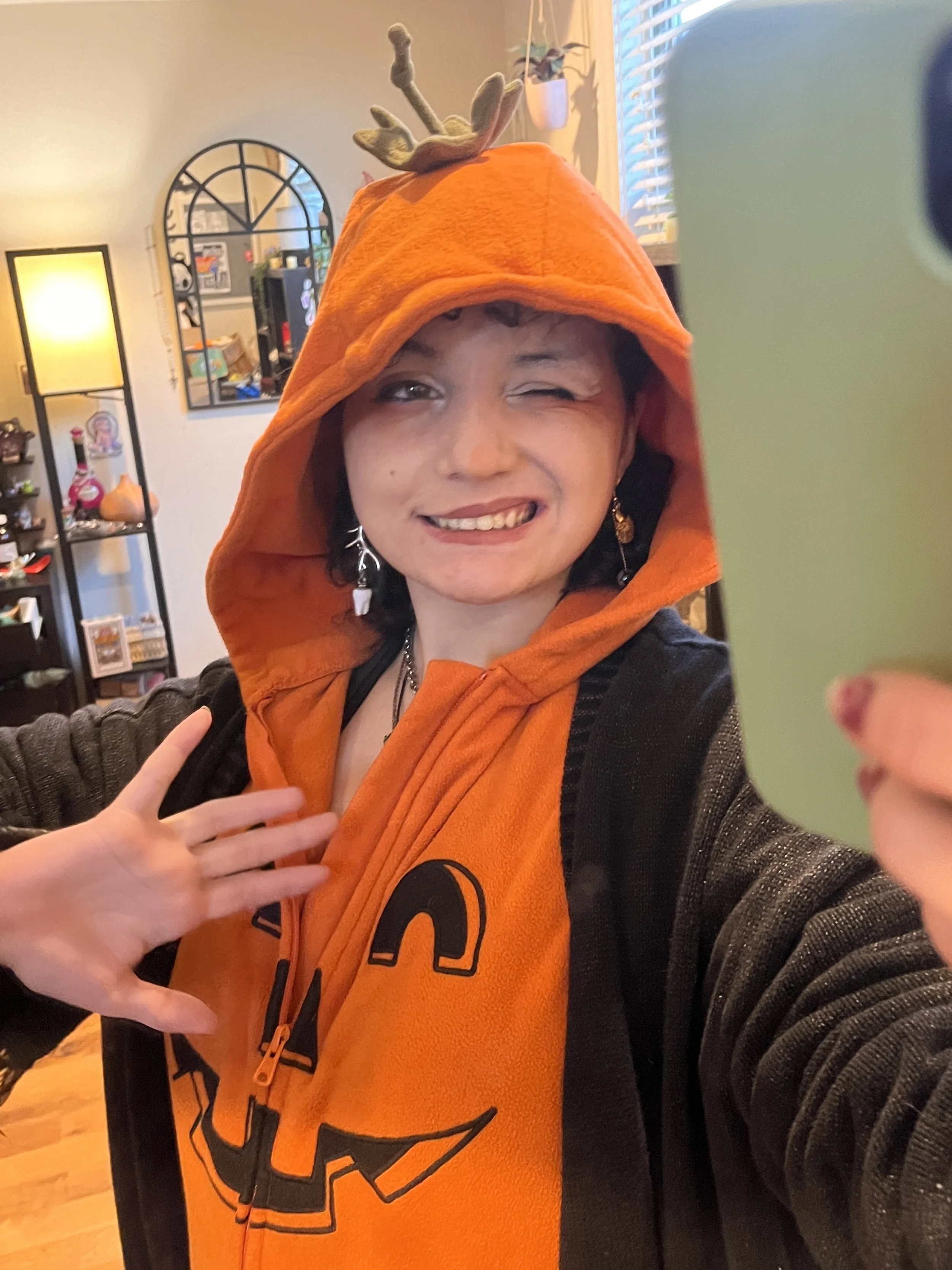Person taking a selfie in costume with an orange hoodie featuring a jack-o'-lantern face and a pumpkin stem on top, winking and smiling in a cozy room.