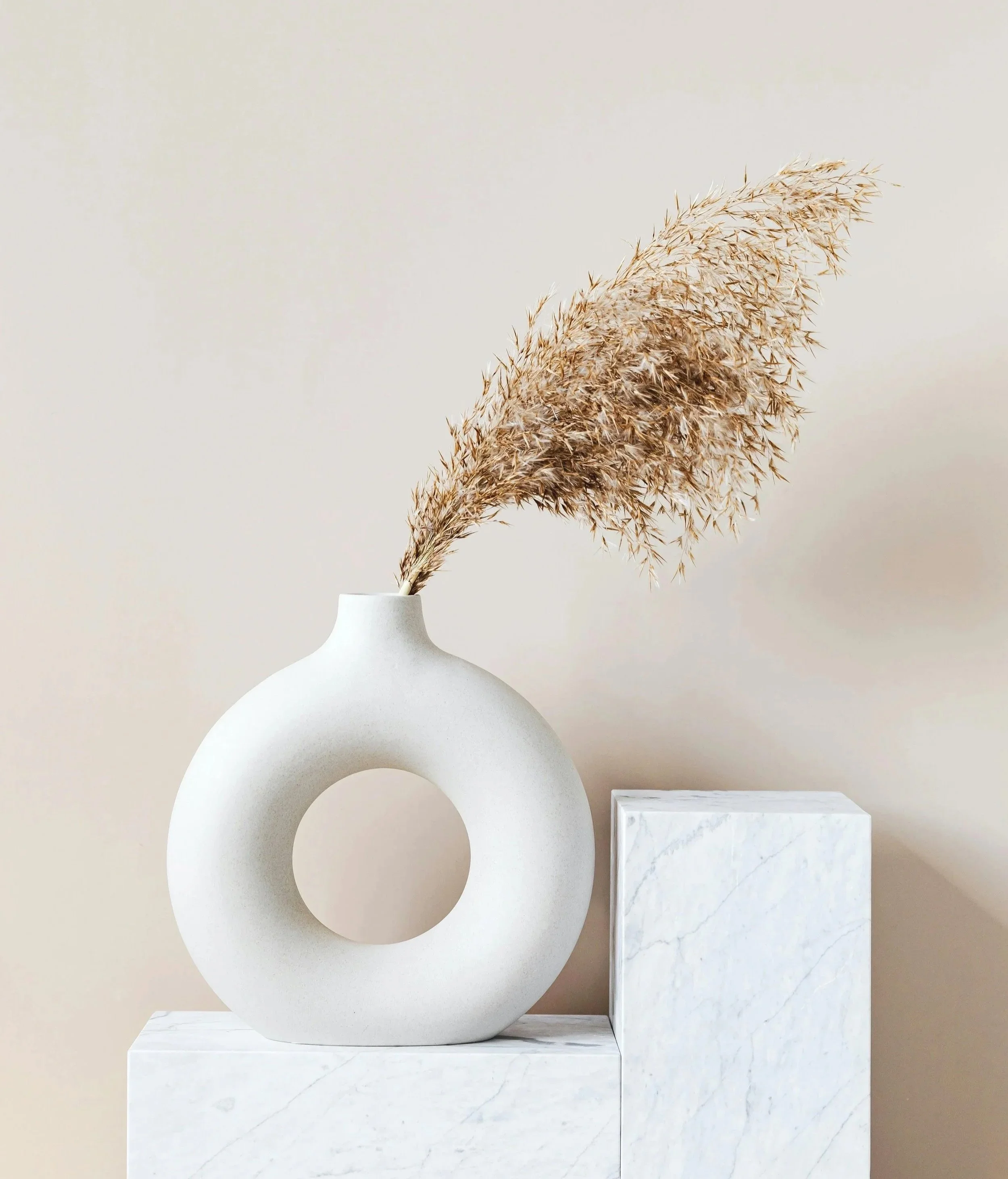A minimalist white ceramic vase with a circular hollow center holding a dried grass stalk, placed on a white marble block against a plain, light-colored wall.