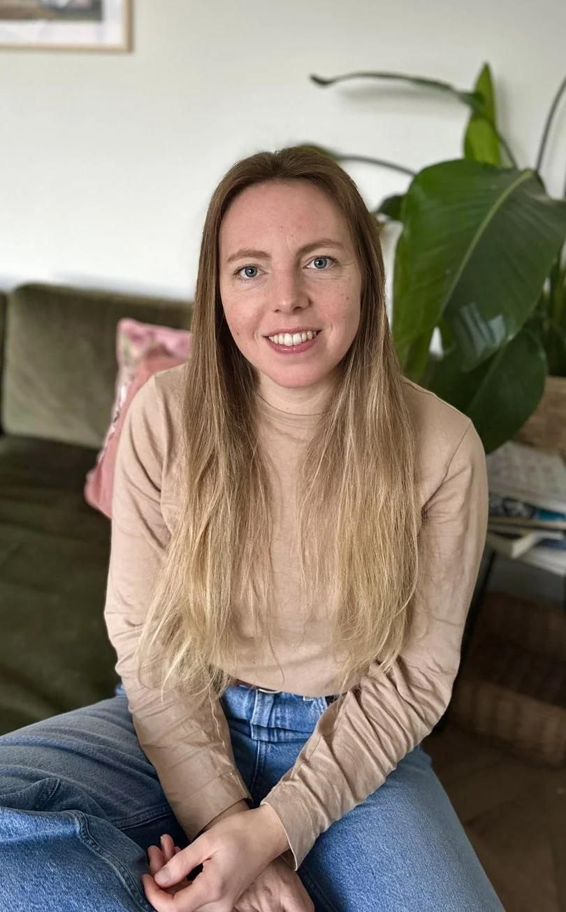 A woman with long, light brown hair, sitting indoors on a wooden piece of furniture with a green couch behind her. There is a pink pillow on the couch and a large leafy plant beside her. The woman is smiling and wearing a beige long-sleeve top and blue jeans.