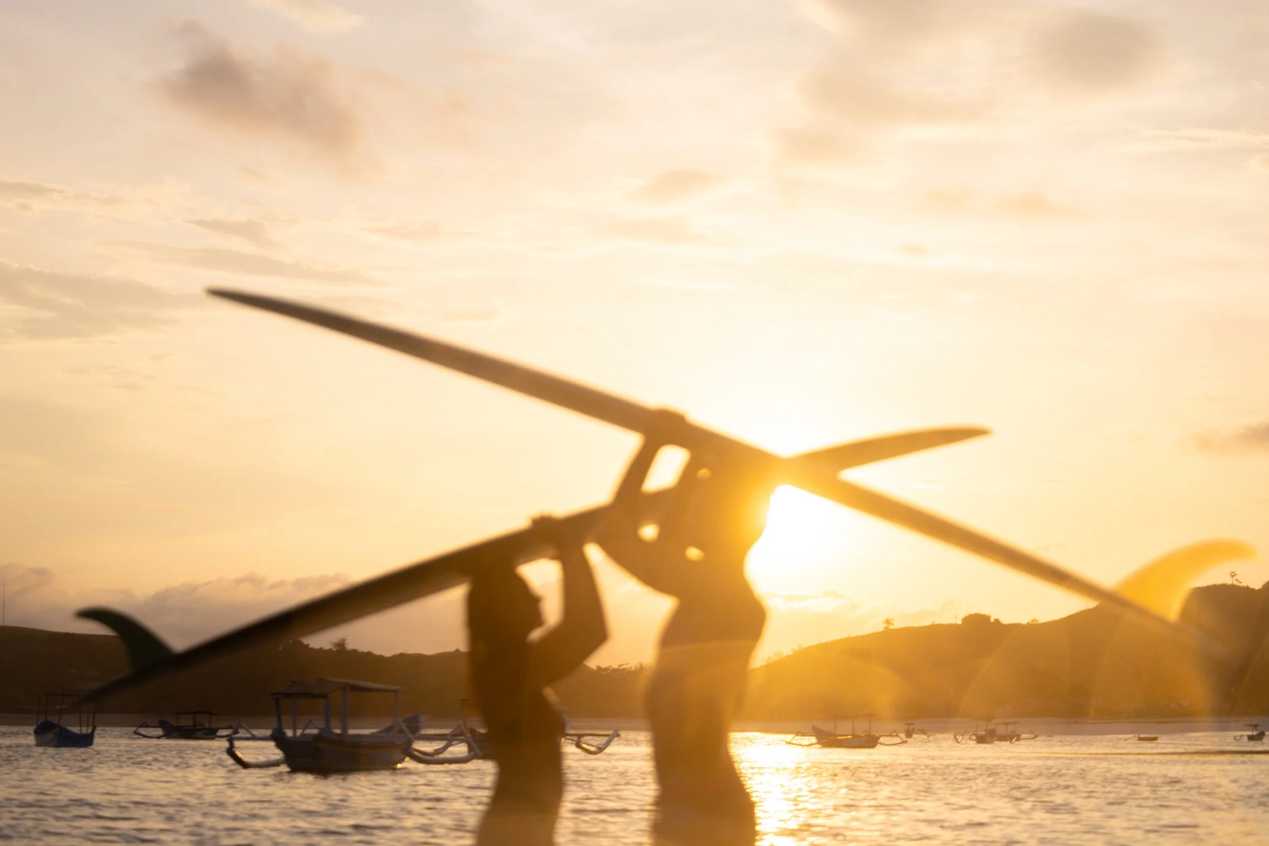 Silhouette of a person carrying a surfboard at sunset over the water with boats and distant hills in the background.