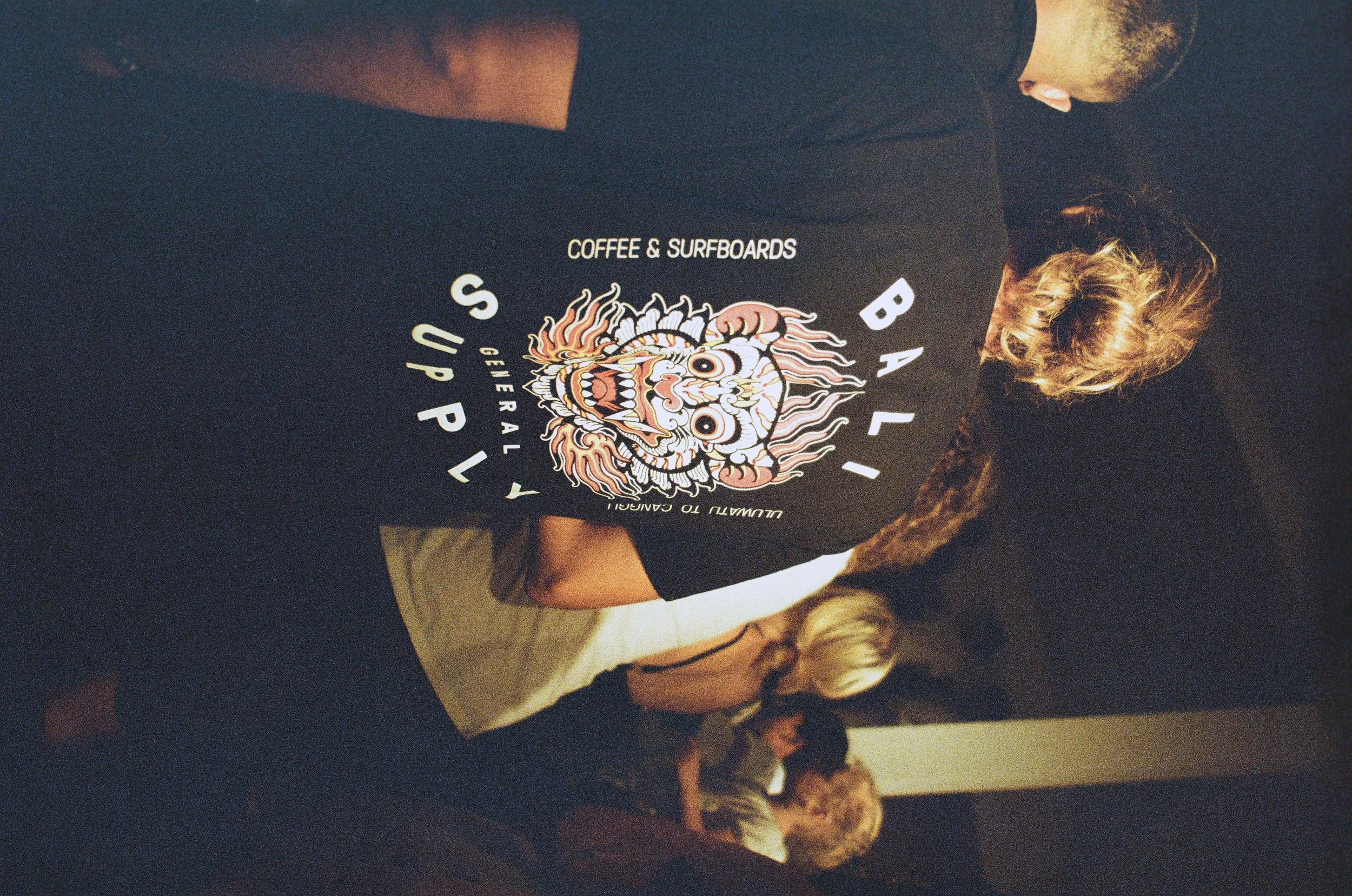 Person wearing a black T-shirt with a colorful lion graphic and the words 'Bali Surf & Coffee' in a dark indoor setting, with other people in the background.