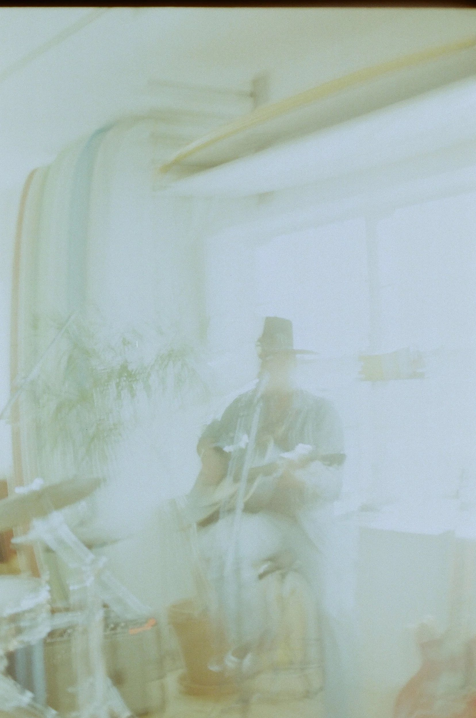 Faded photo of Donavon Frankenreiter on a chair, playing a guitar, near a window in Adam Mar Surf Shop