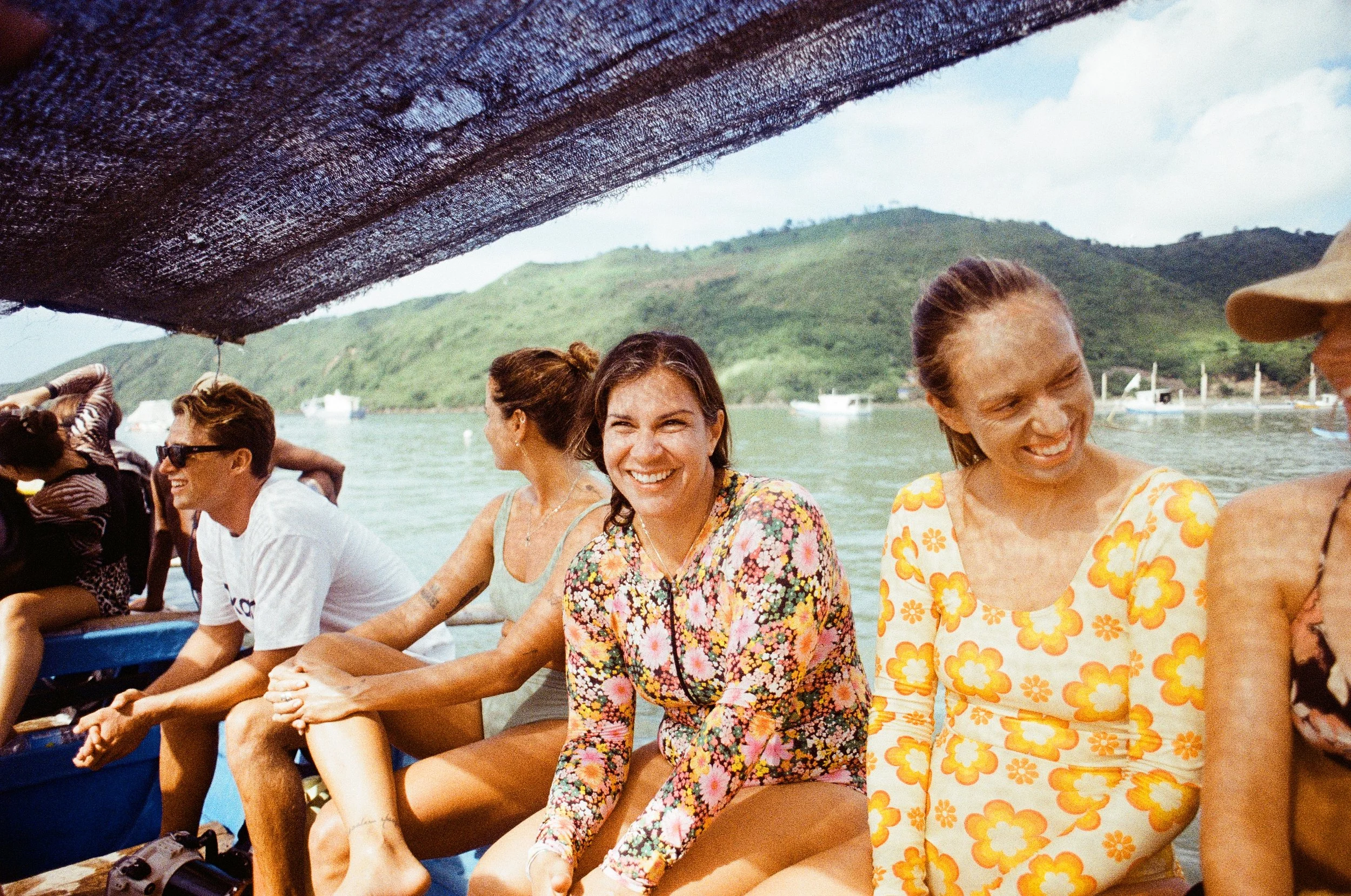 Group of smiling women and a man sitting on a boat with a scenic water and green hills background.