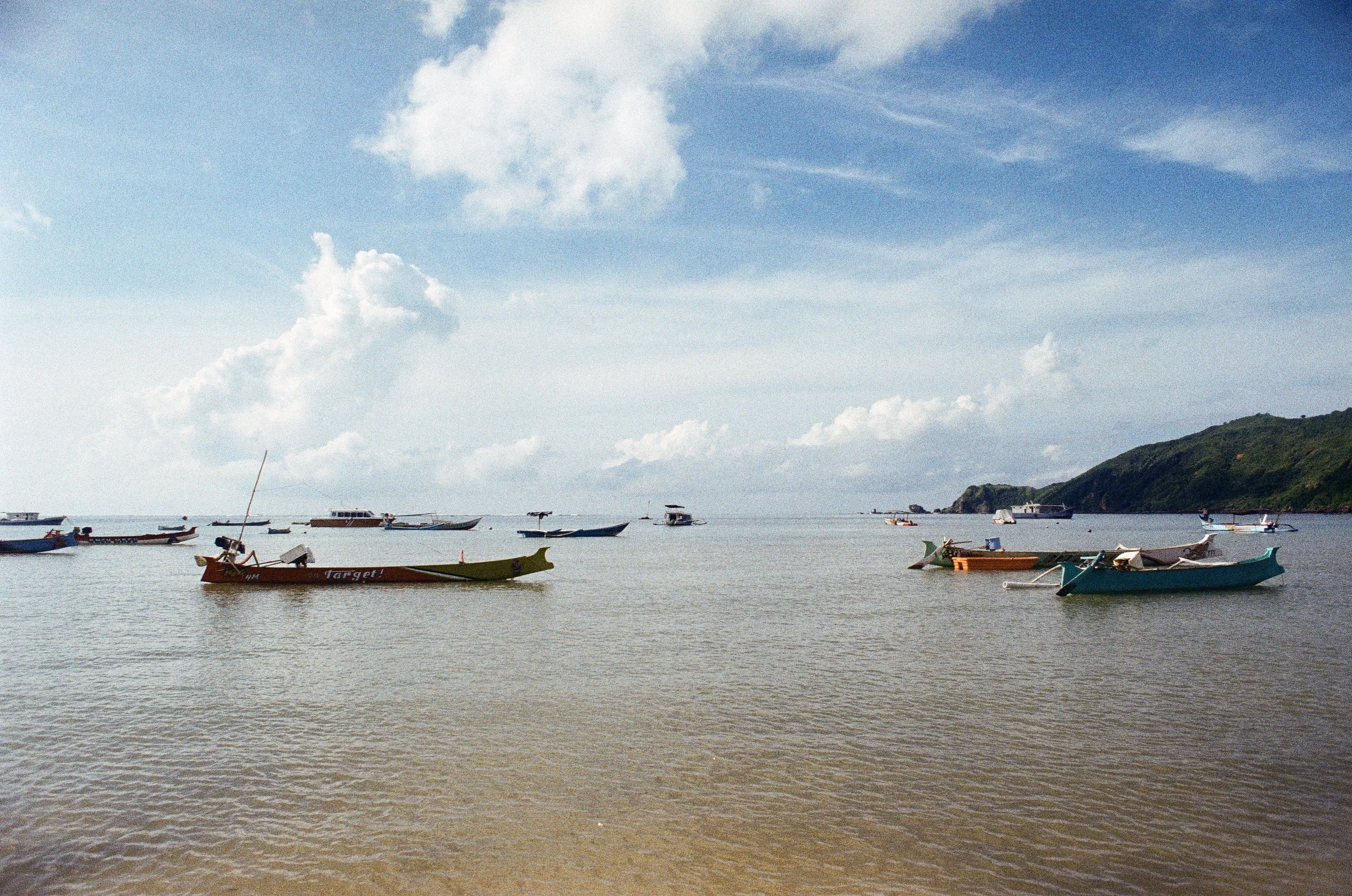 Several small boats floating on calm water near a sandy shoreline, with a island or hill covered in greenery in the background and a partly cloudy sky overhead.
