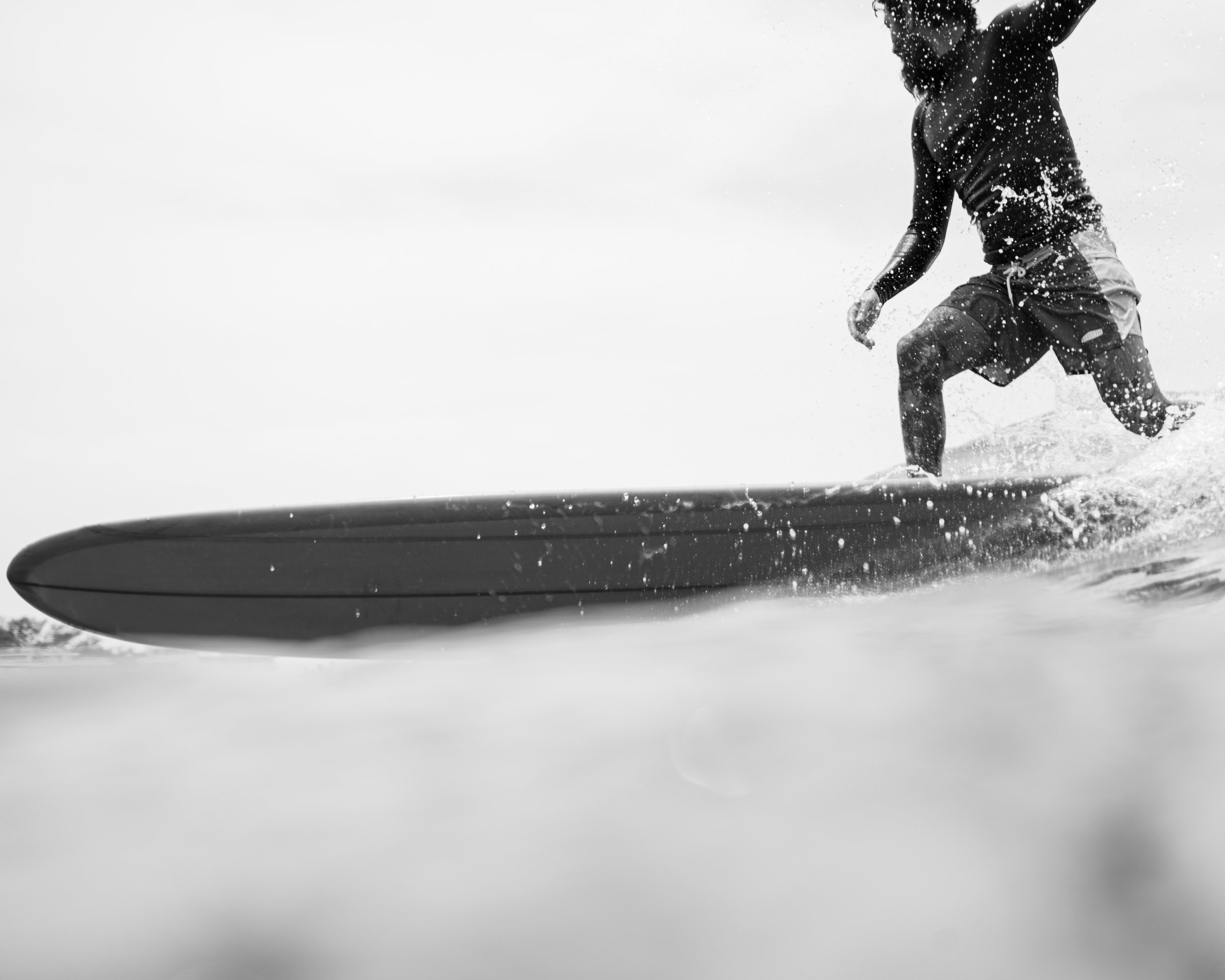 Black and white photo of a man standing on his surfboard, surfing on water with spray and waves around him.