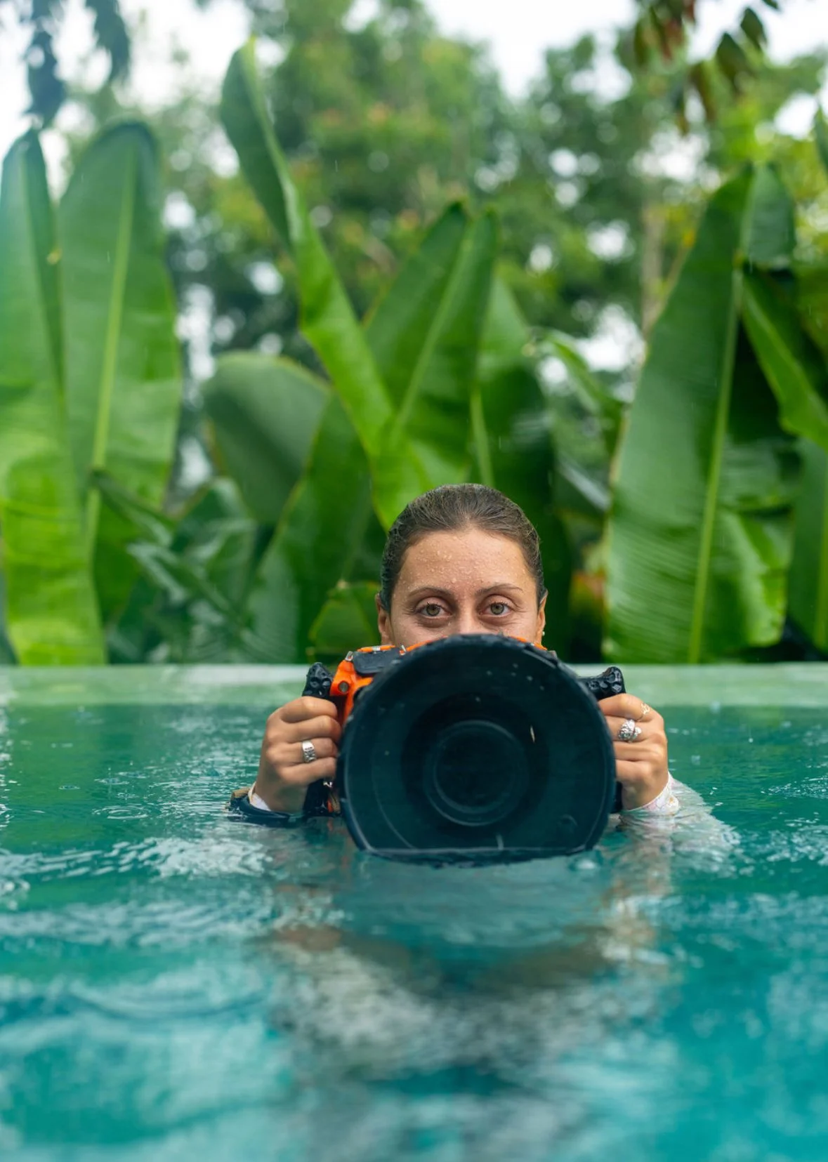 A woman holding a camera in a pool, with lush green plants in the background.
