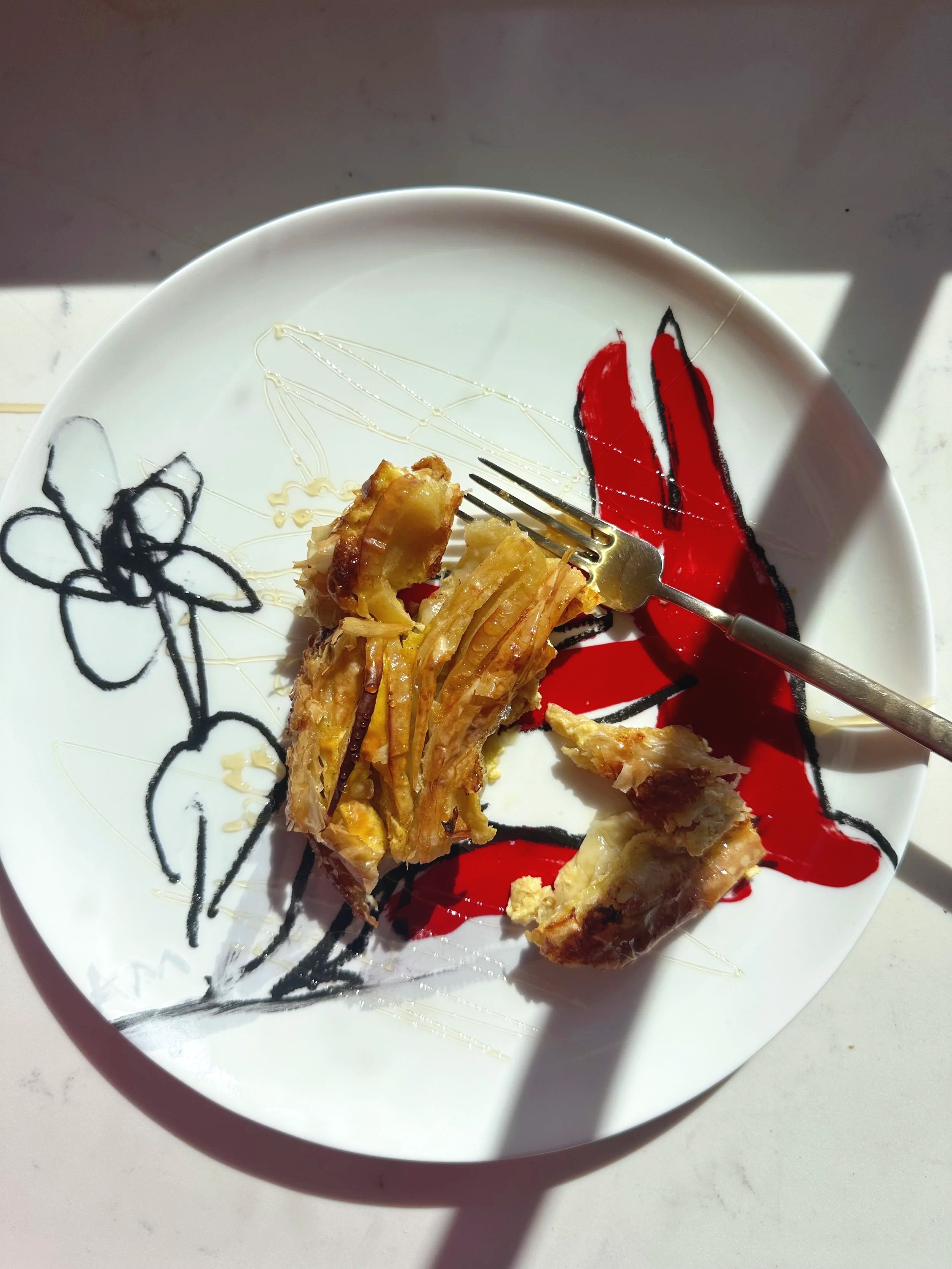 A partially eaten piece of chicken on a decorative plate with black and red floral designs, with a fork resting on the plate, and direct sunlight casting shadows.