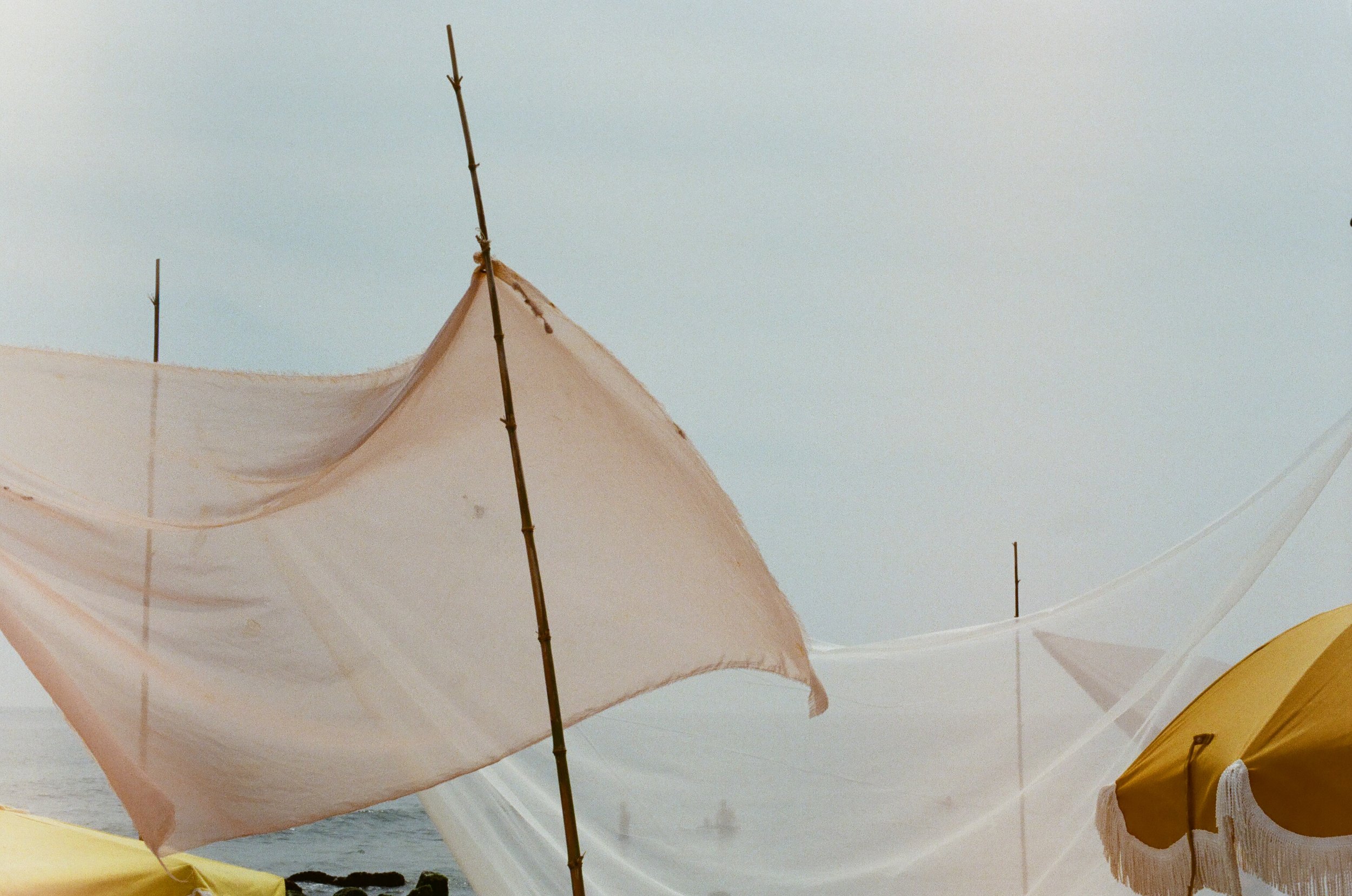 Beach scene with pastel-colored and yellow umbrellas and fabric drapes, over the ocean with a cloudy sky in the background.