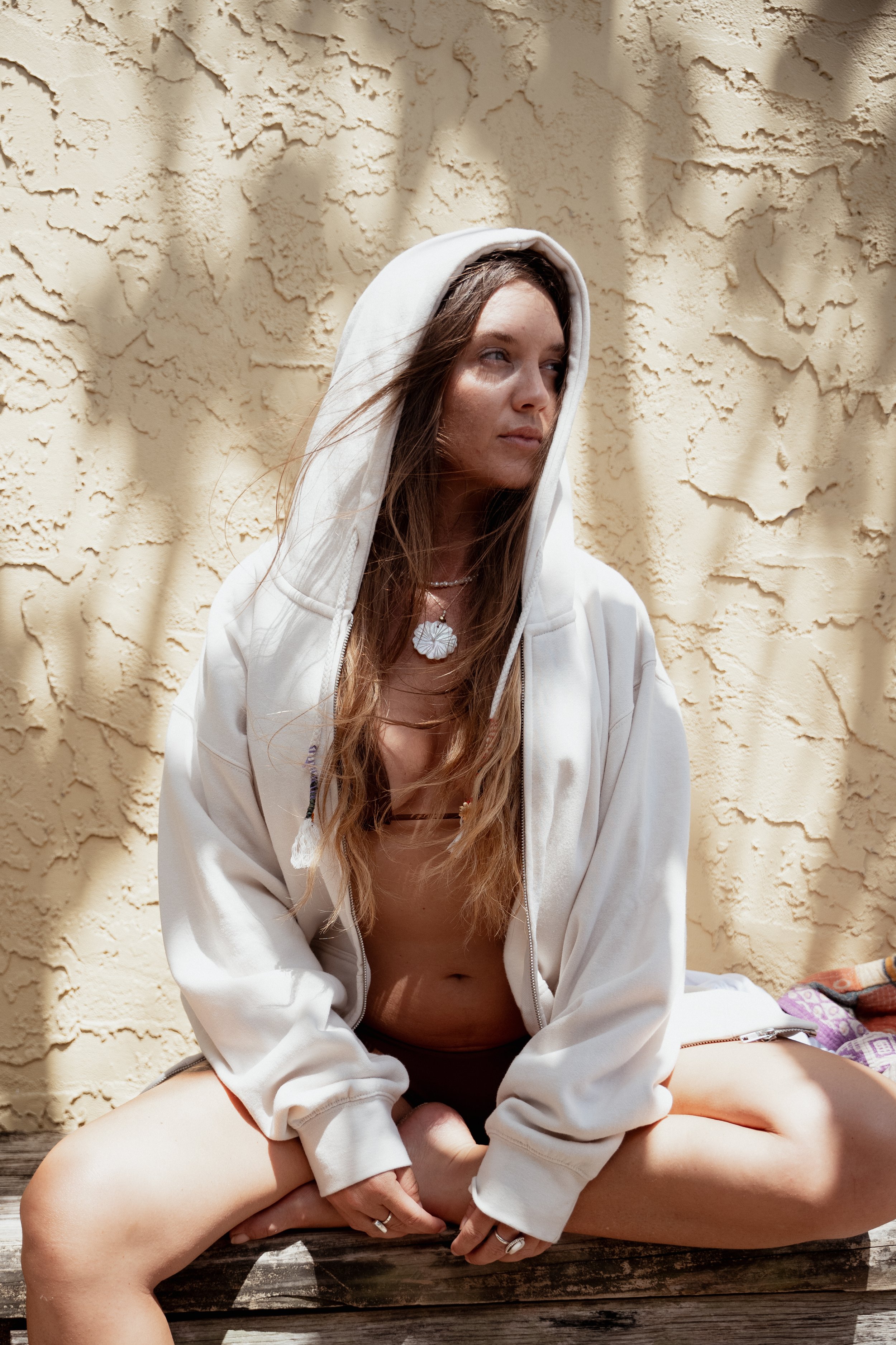 A young woman with light skin and long brown hair, wearing a hoodie with the hood up, sitting cross-legged on a wooden surface in front of a textured beige wall. She looks to the side with a neutral expression, and she is accessorized with necklaces 