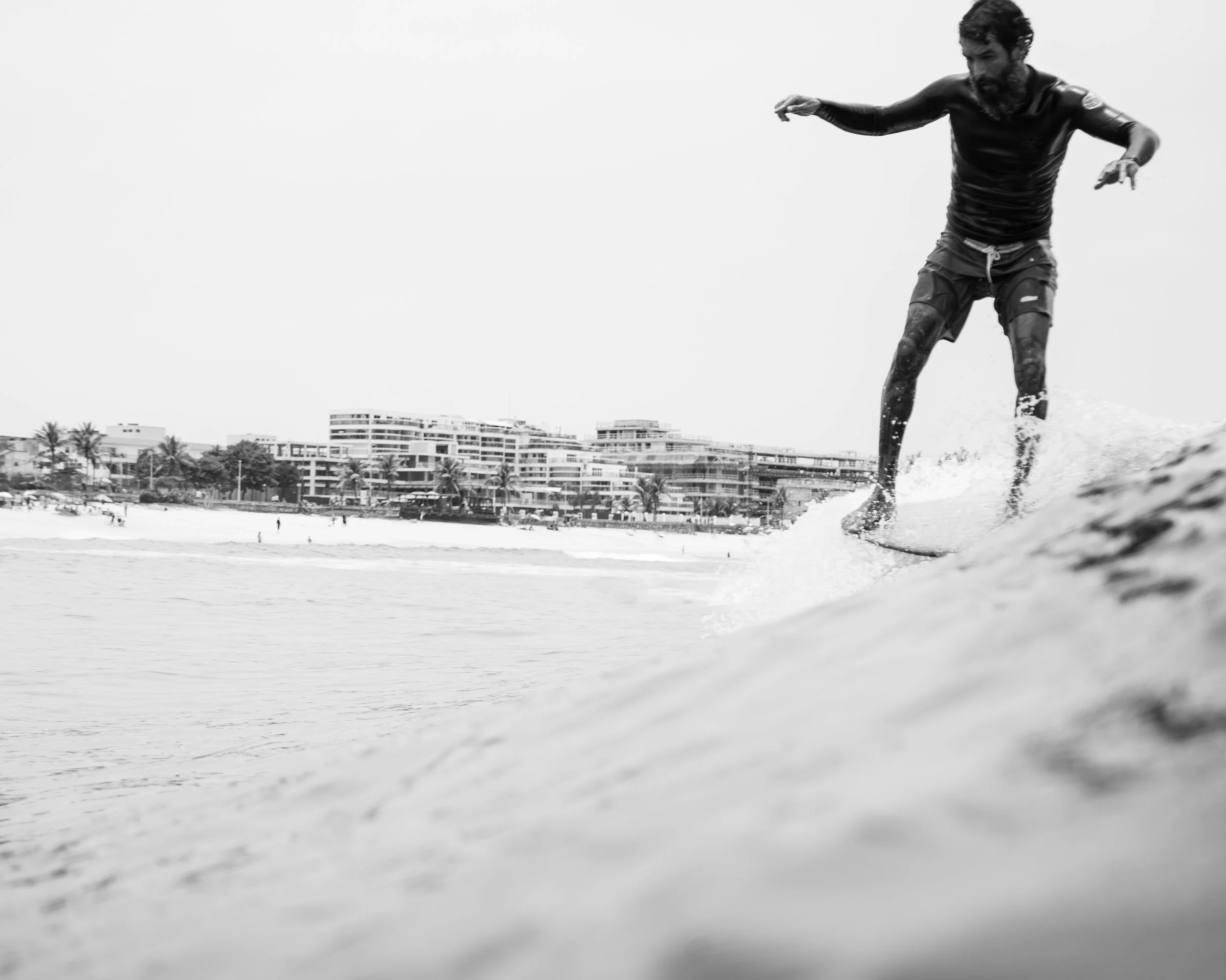Man surfing on a wave at the beach, black and white photo with city buildings and palm trees in the background.