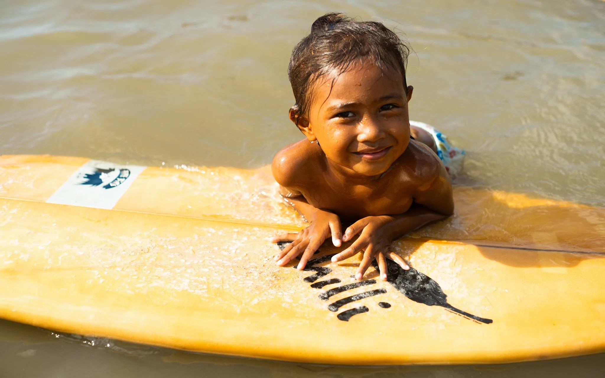 Young boy with wet hair smiling as he lies on a yellow surfboard in the water.