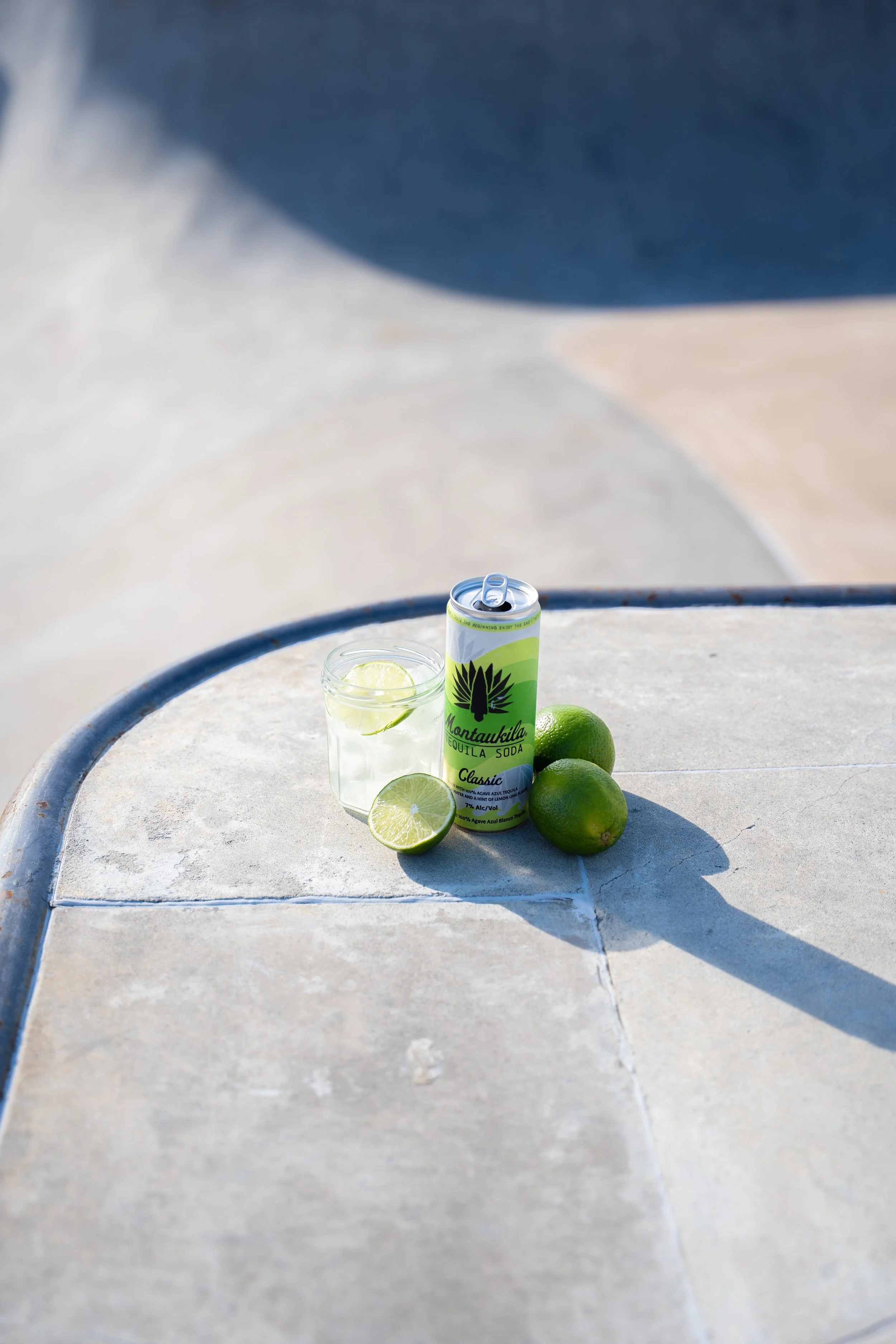 A can of Montaukila Soda Classic, a glass with lime slices and ice, and three whole limes on a concrete surface in sunlight.
