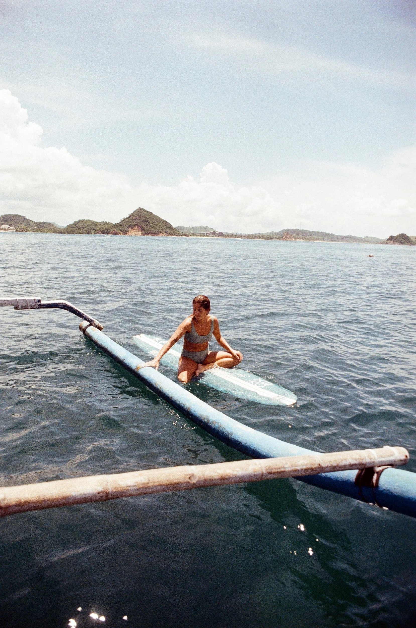 A woman in a swimsuit sitting on a surfboard in the water near an outrigger boat.