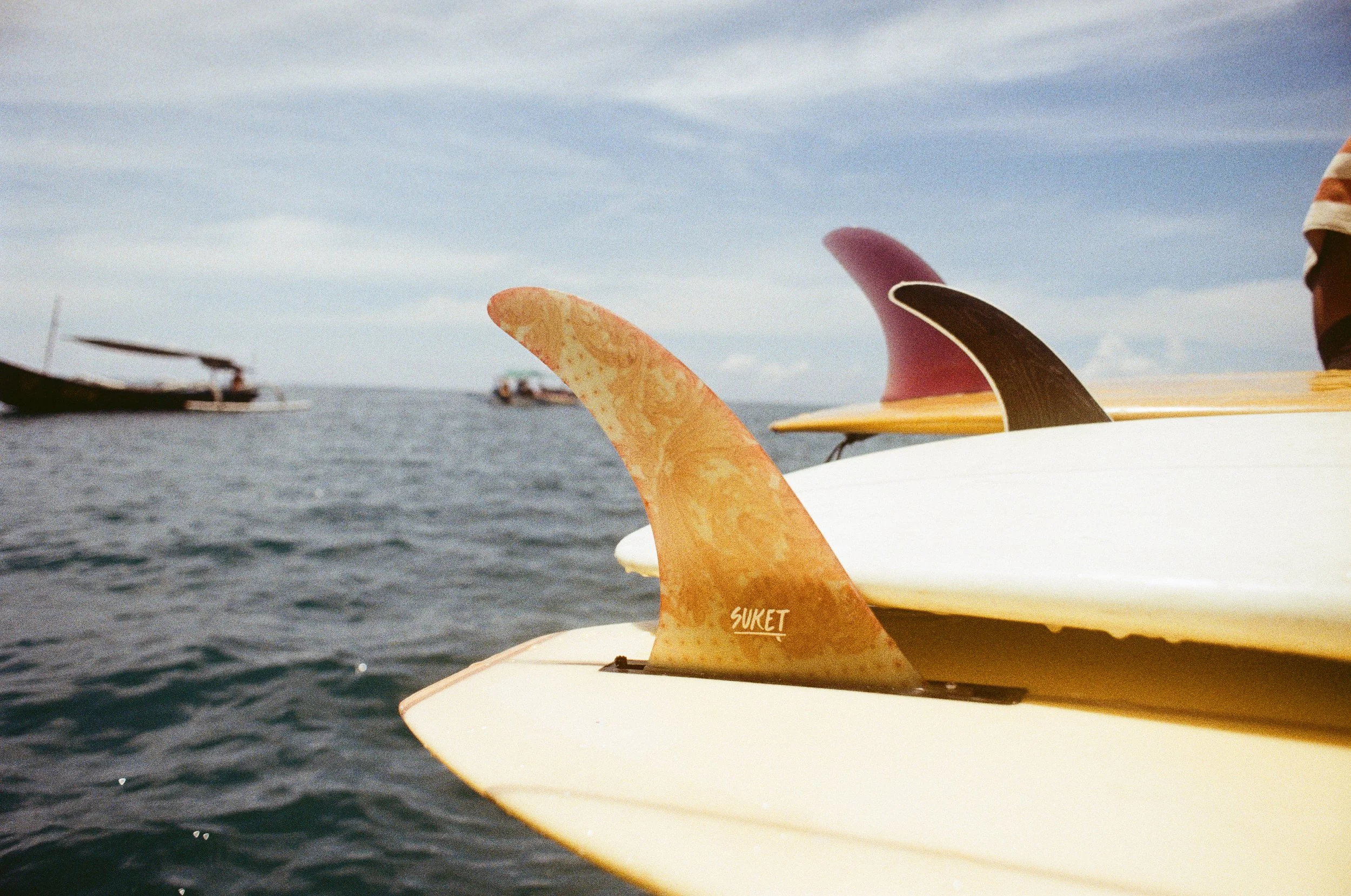 Close-up of three surfboard fins on a yellow surfboard, with boats on the water and a cloudy sky in the background.