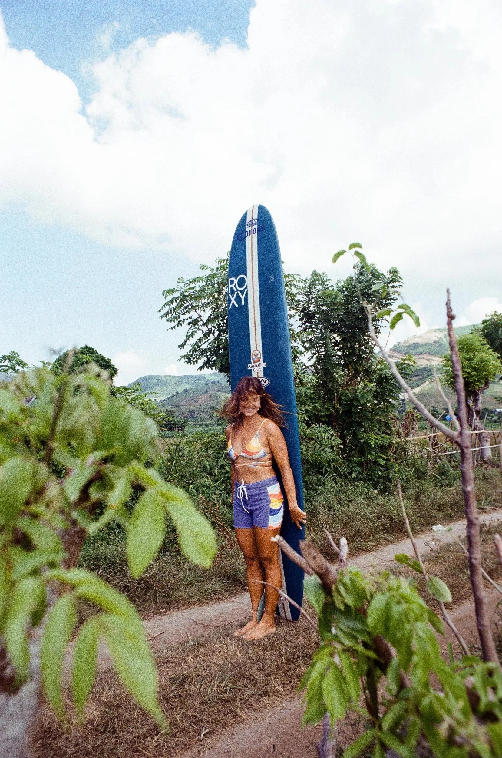 A woman in a colorful bikini and shorts standing barefoot on a dirt path, next to a large blue surfboard, smiling and posing outdoors with green trees and mountains in the background.