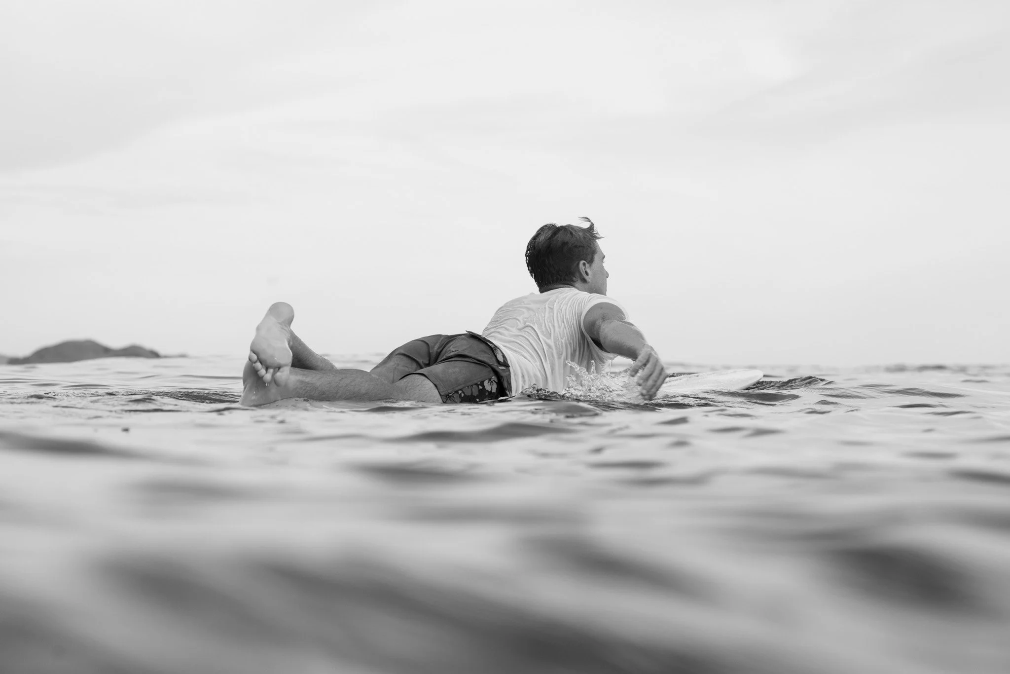 A man paddling to a wave in the ocean, lying prone on a surfboard, with a distant landmass in the background.