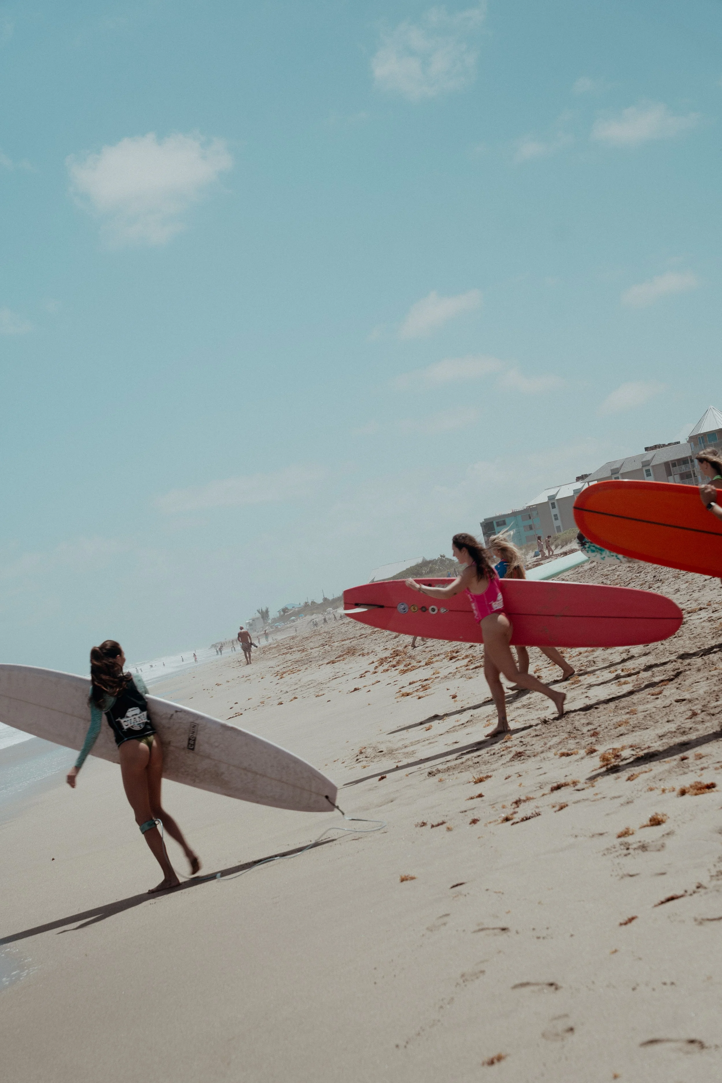 People walking with surfboards on a sandy beach under a partly cloudy sky.