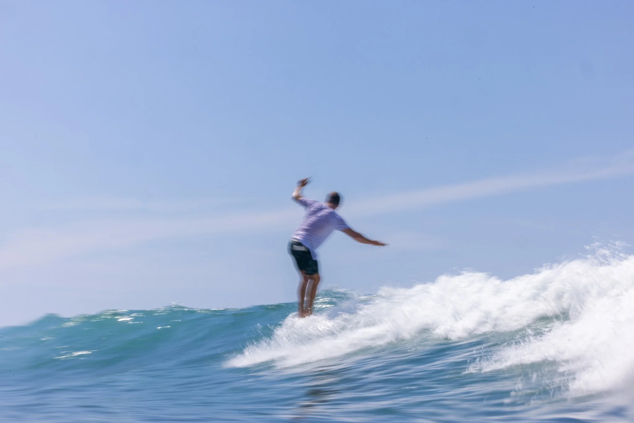 Man in a white shirt and black shorts riding a wave on a surfboard under a clear blue sky.