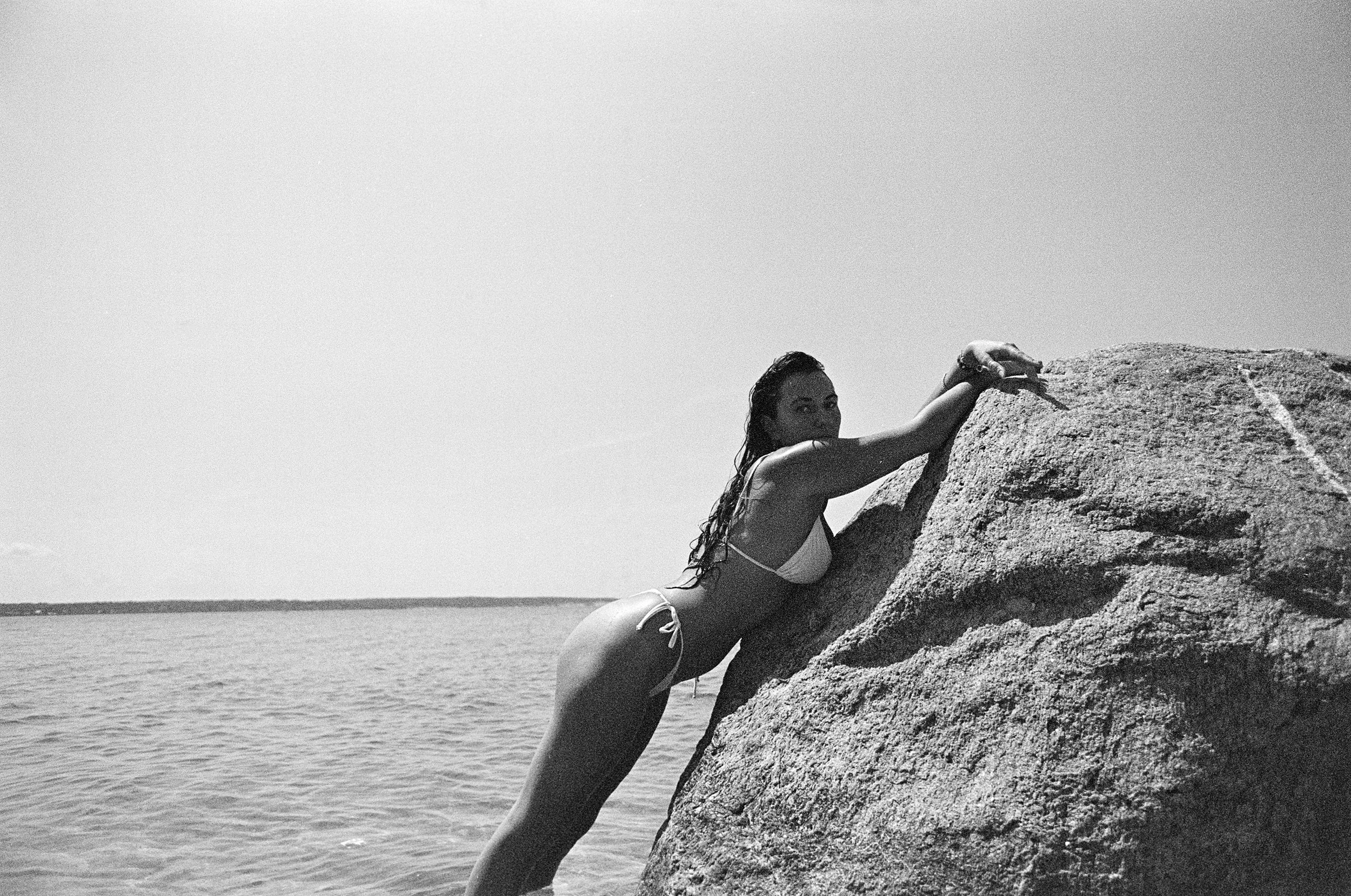 A woman in a bikini leaning on a large rock by the water, looking back over her shoulder.